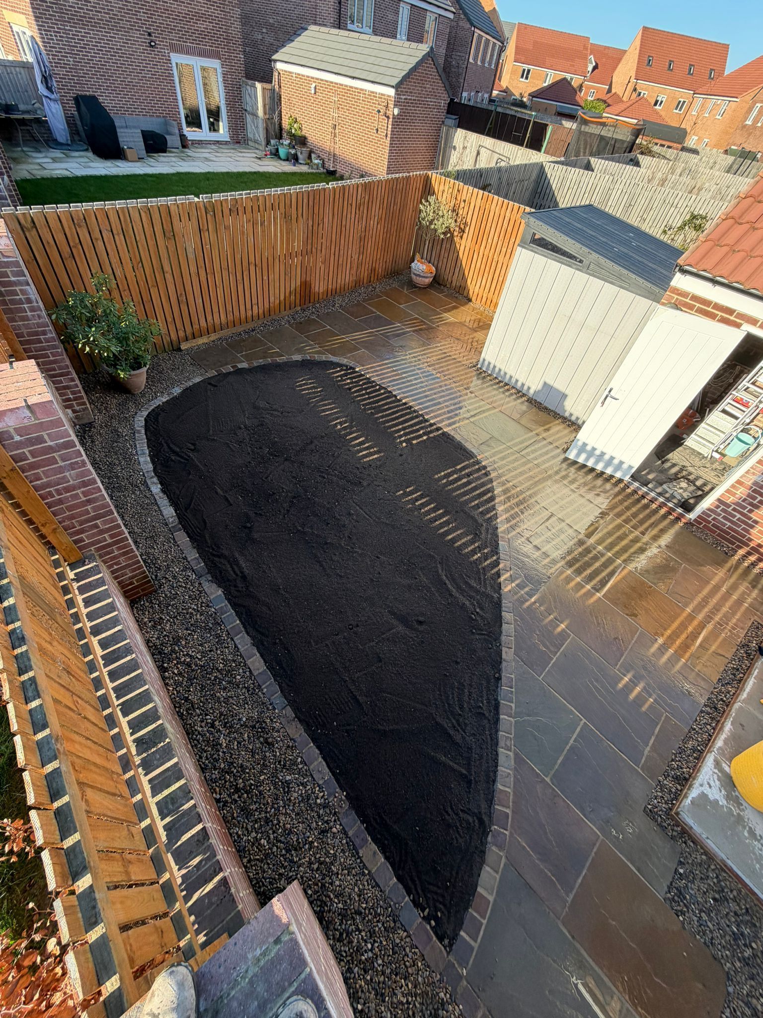 Backyard with a large dark pit beside a brick wall, viewed from above in bright sunlight