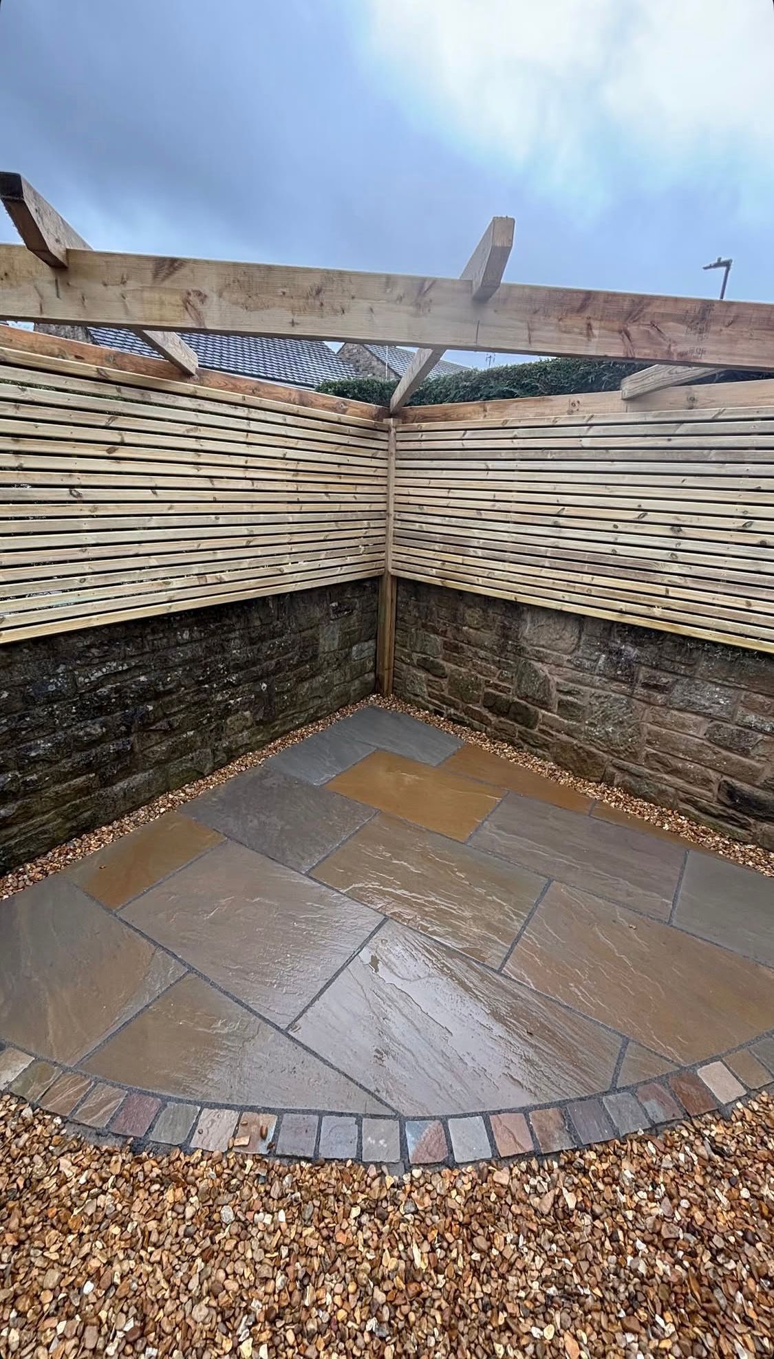 Empty corner patio with woven fence, stone paving, and gravel under an open sky.