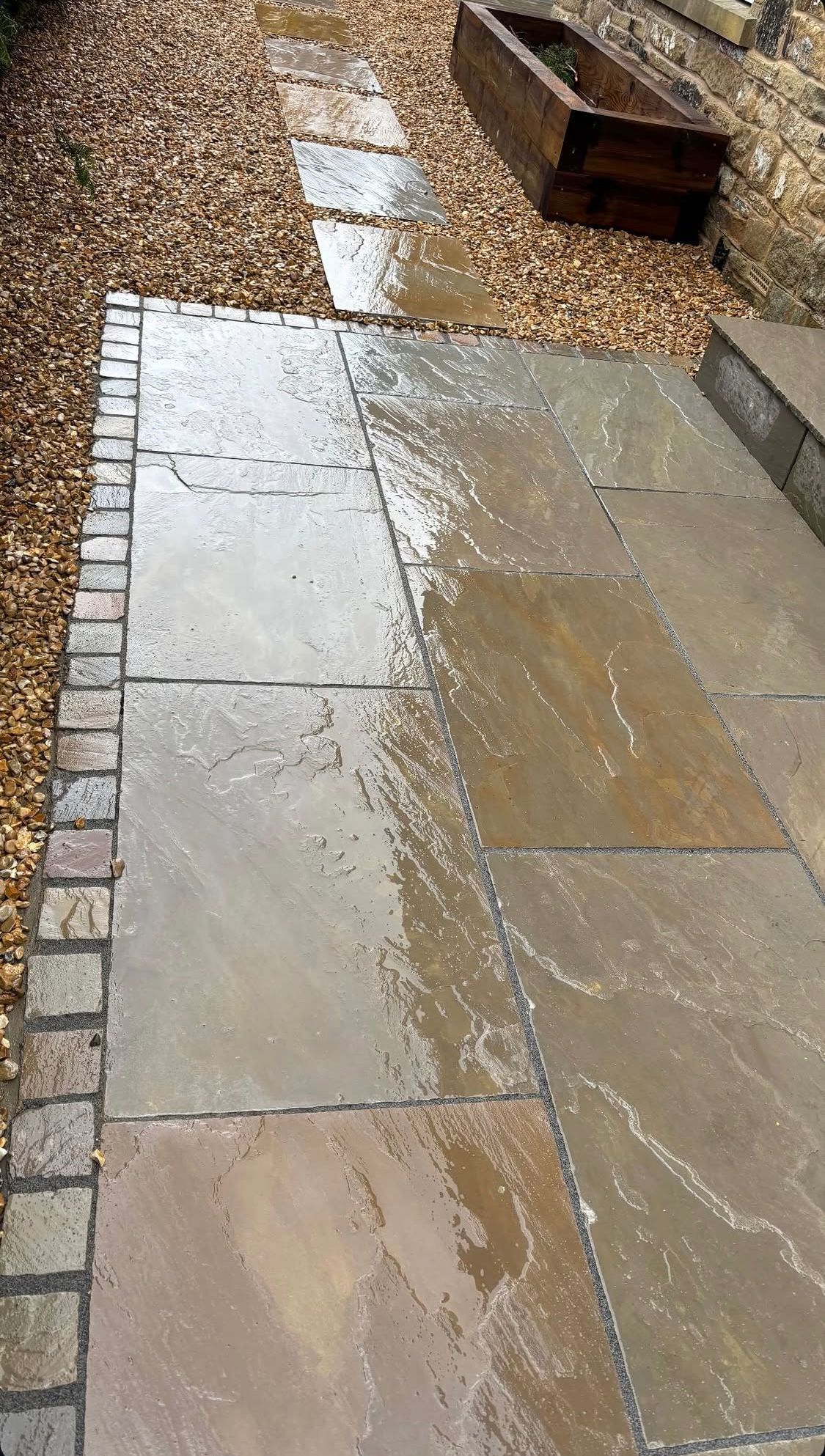 Wet stone patio with rainwater pooling beside gravel and a wooden bench