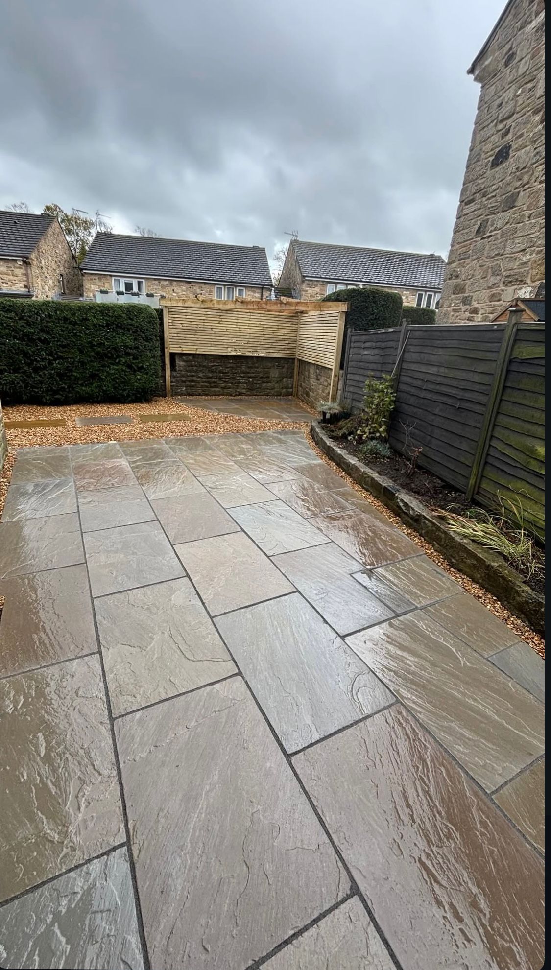 Wet stone patio beside a hedge and fence, with a cloudy sky and houses in the background.
