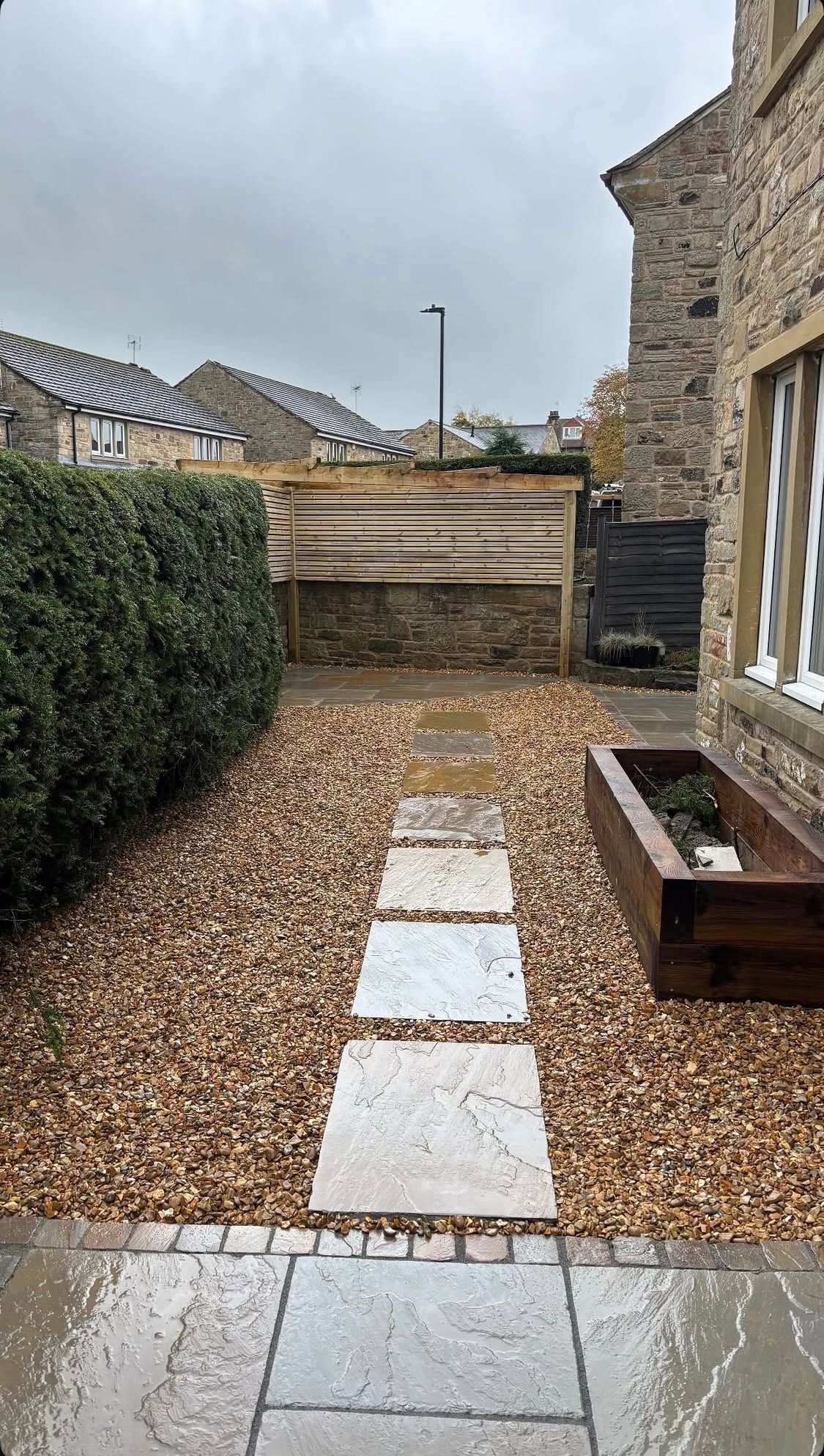 Narrow gravel garden path beside a stone house, with stepping stones, hedge, and wooden bench under a cloudy sky