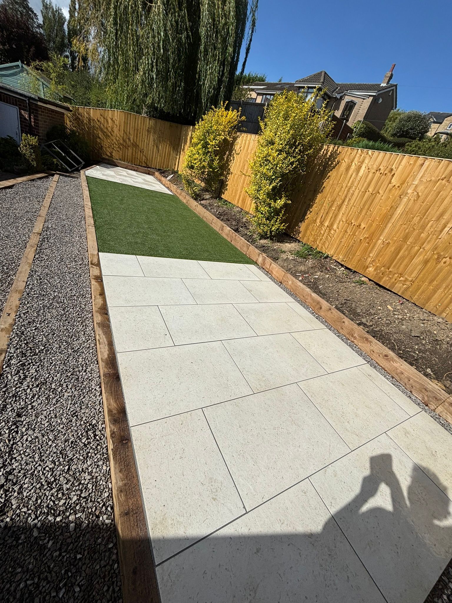 Backyard patio with gravel, concrete pavers, a small grass strip, and a wooden fence in bright sunlight