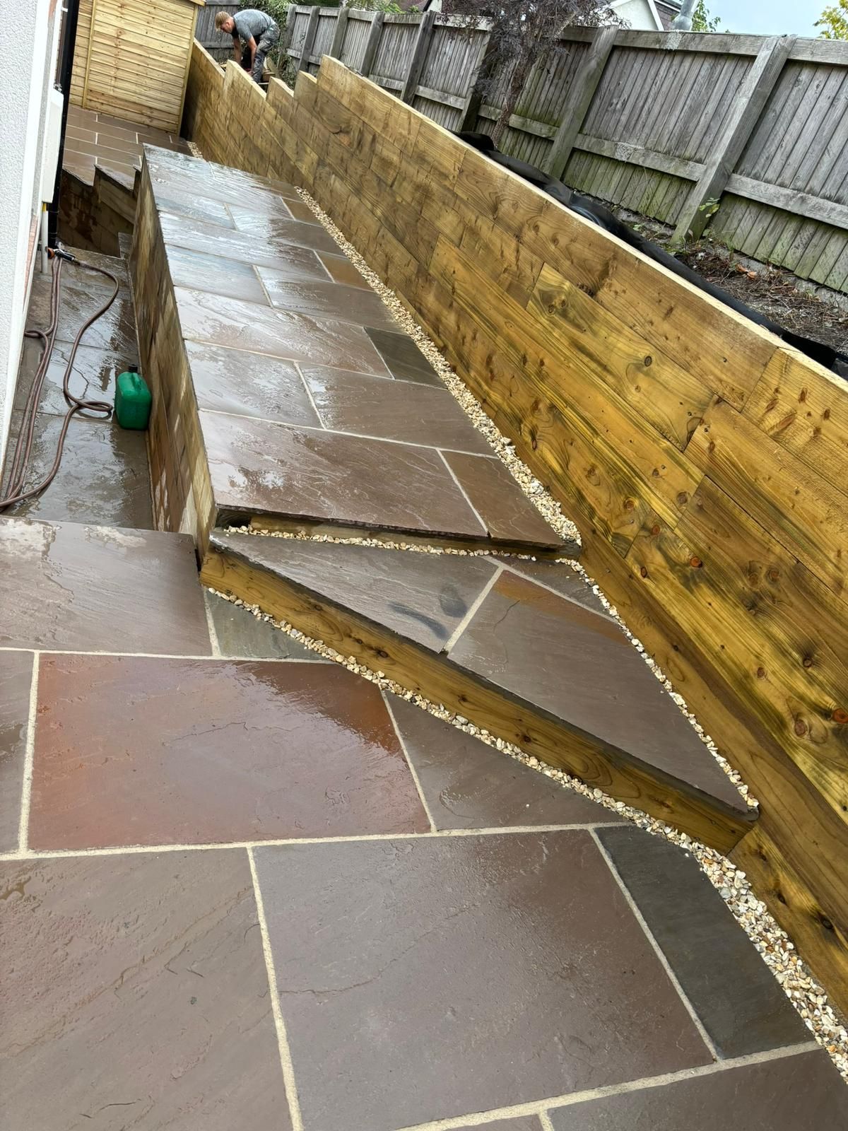 Sloped wet patio slabs beside a wooden fence, with a damaged raised section causing a trip hazard.
