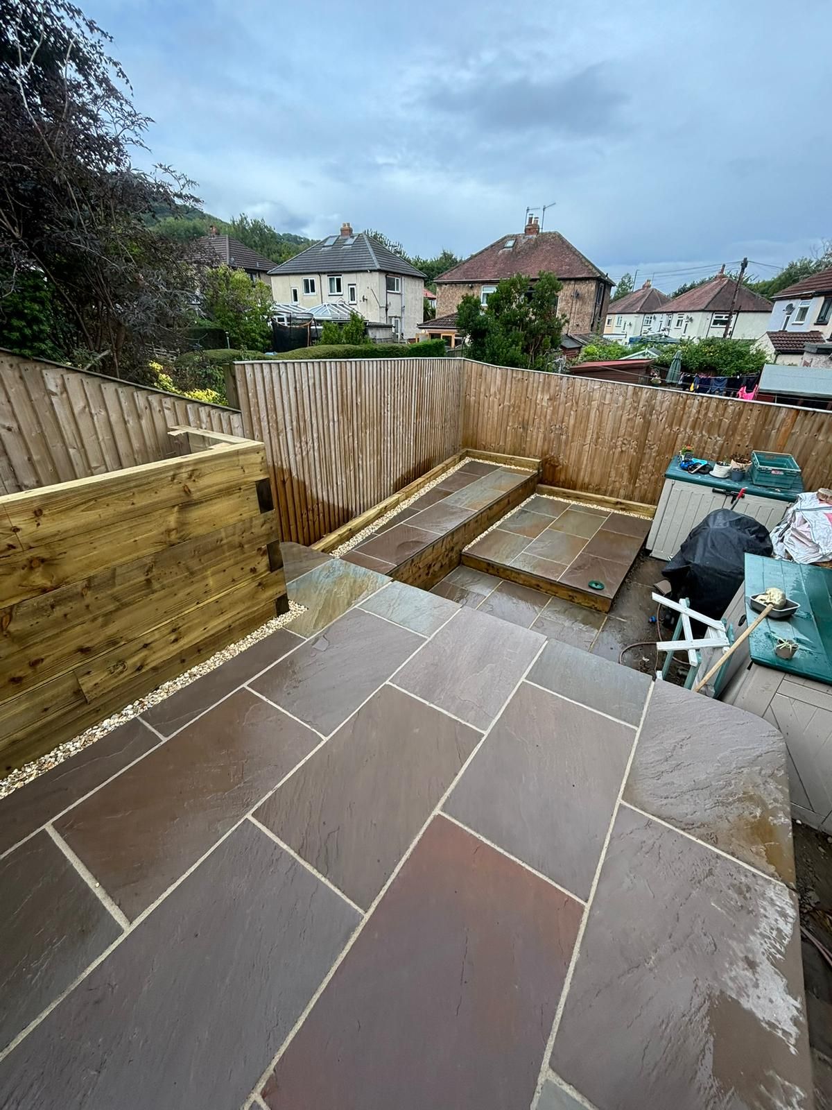 Backyard patio with large stone slabs, wooden fencing, and a small raised garden bed under cloudy sky