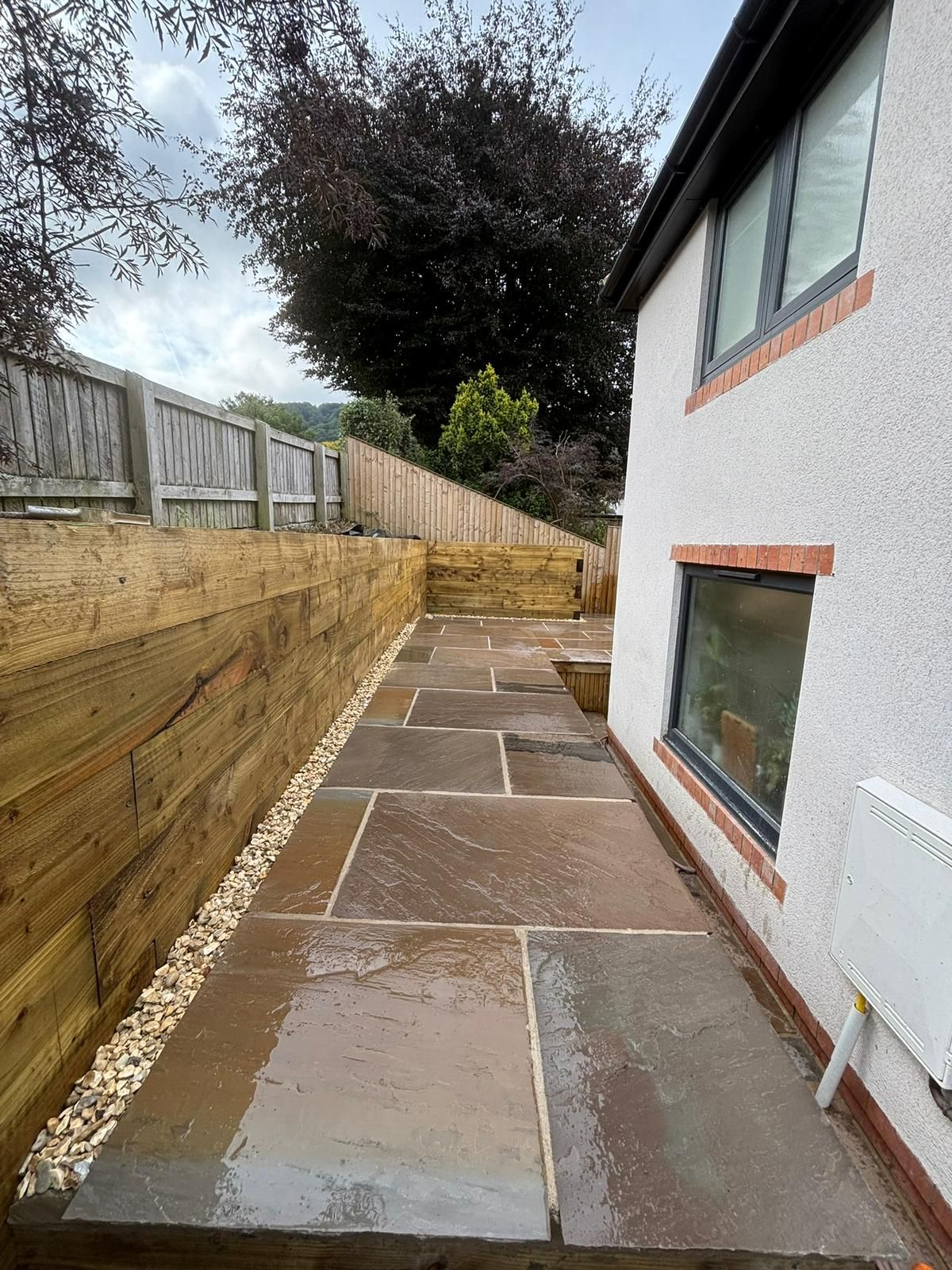 Narrow side yard with stone paving beside a white house and wooden fence on a cloudy day