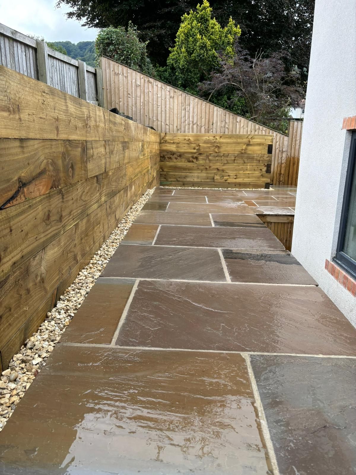 Wet stone patio path beside wooden fence and house wall leading to a raised deck area