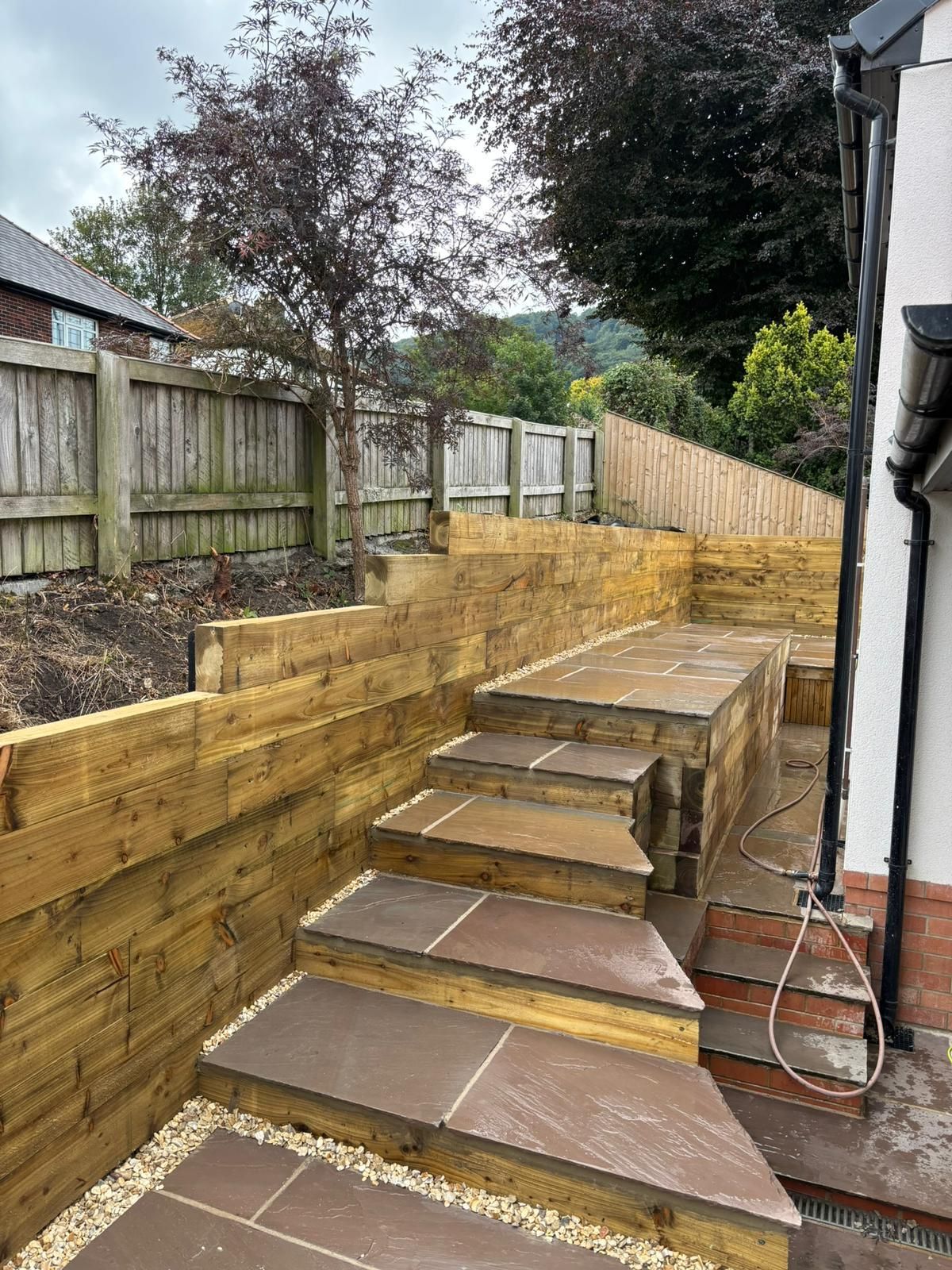 Wooden stepped garden retaining wall and stairs beside a house, with fencing and trees along a sloped yard.