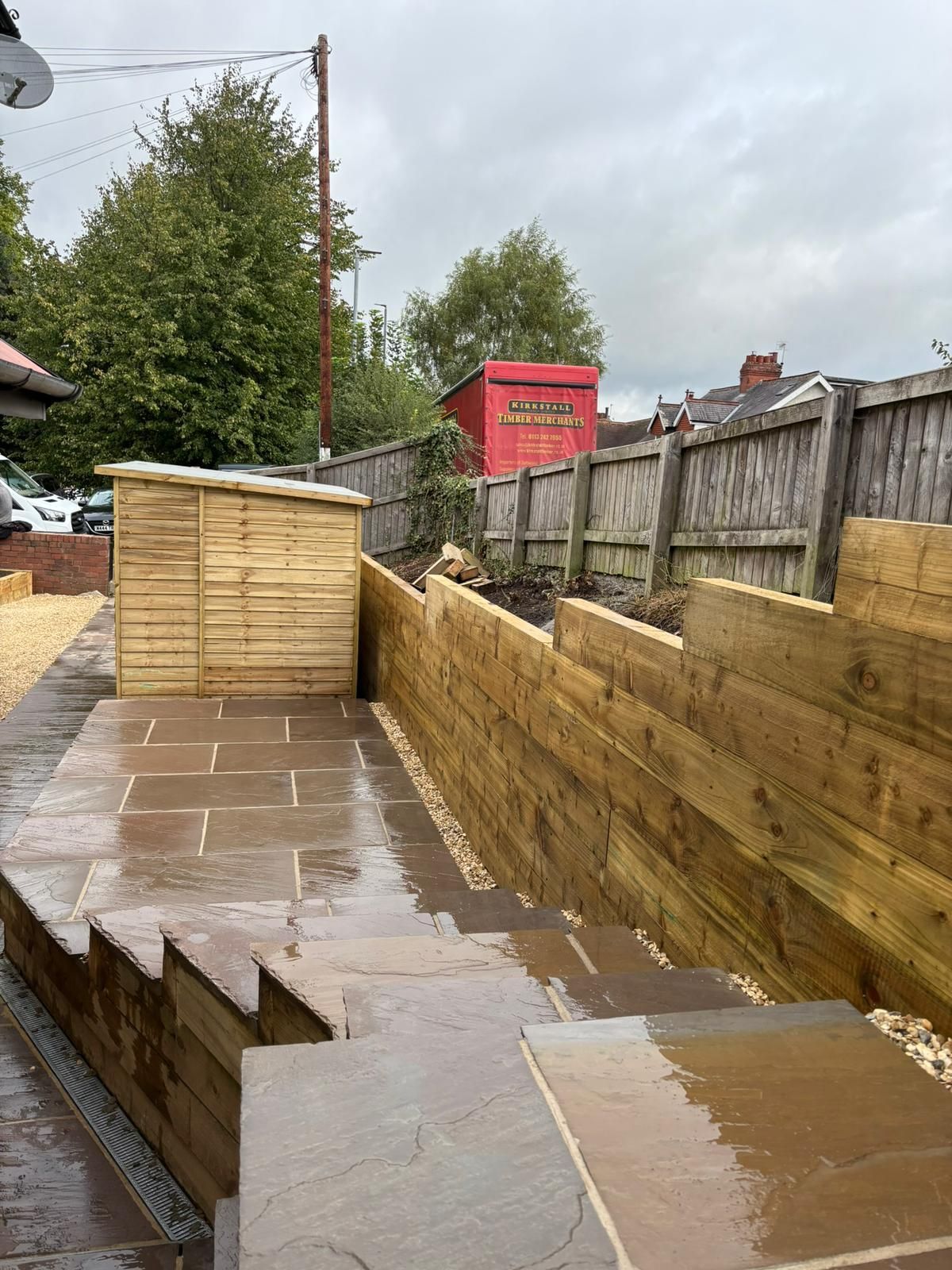 Wet paved backyard with wooden retaining walls, shed, and fence on a cloudy day