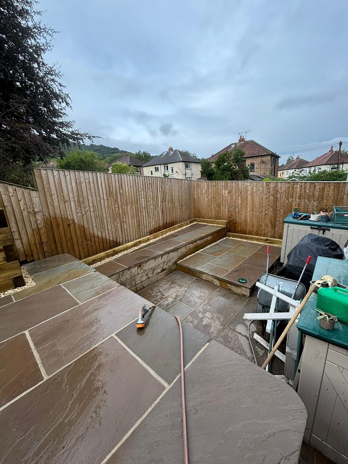 Backyard patio under construction with paving slabs, sand, tools, and a wooden fence.