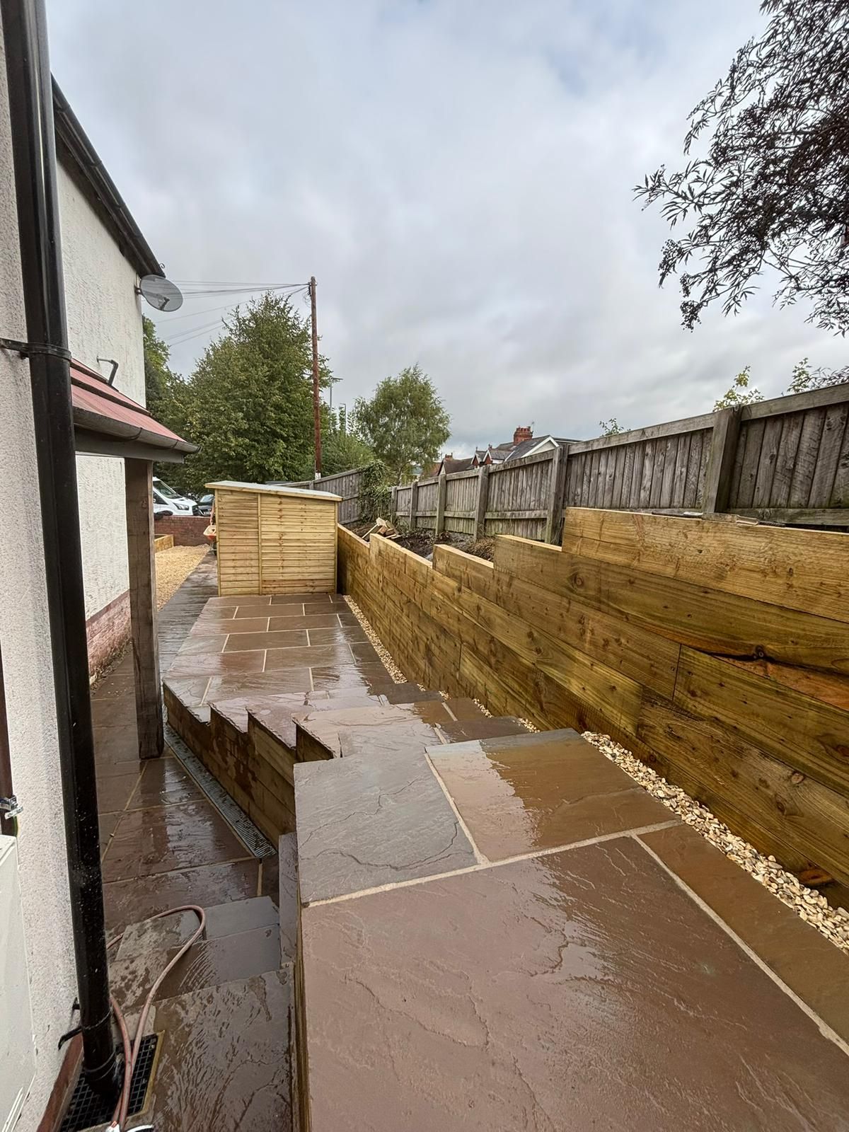 Backyard patio with wet concrete walkway, wooden retaining walls, and a fence on a rainy day
