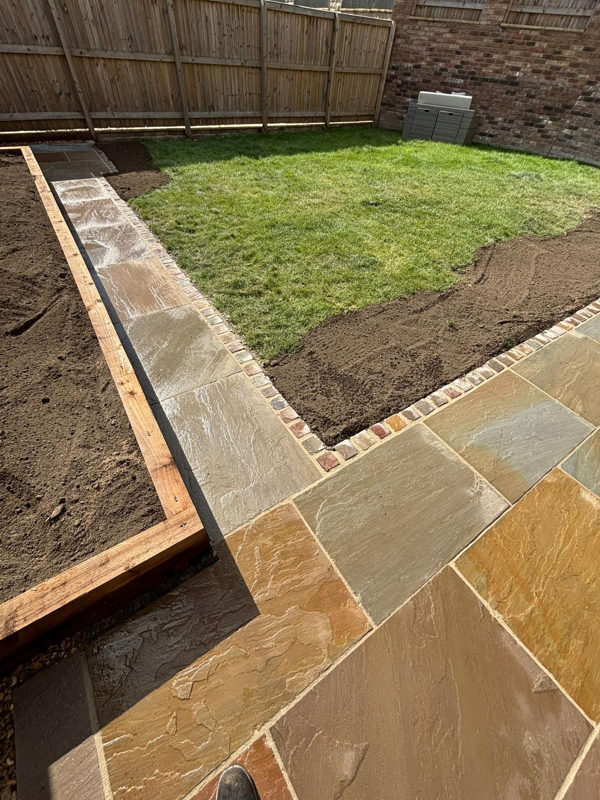 Backyard patio with stone pavers, grass center, and a raised soil bed beside a wooden fence.