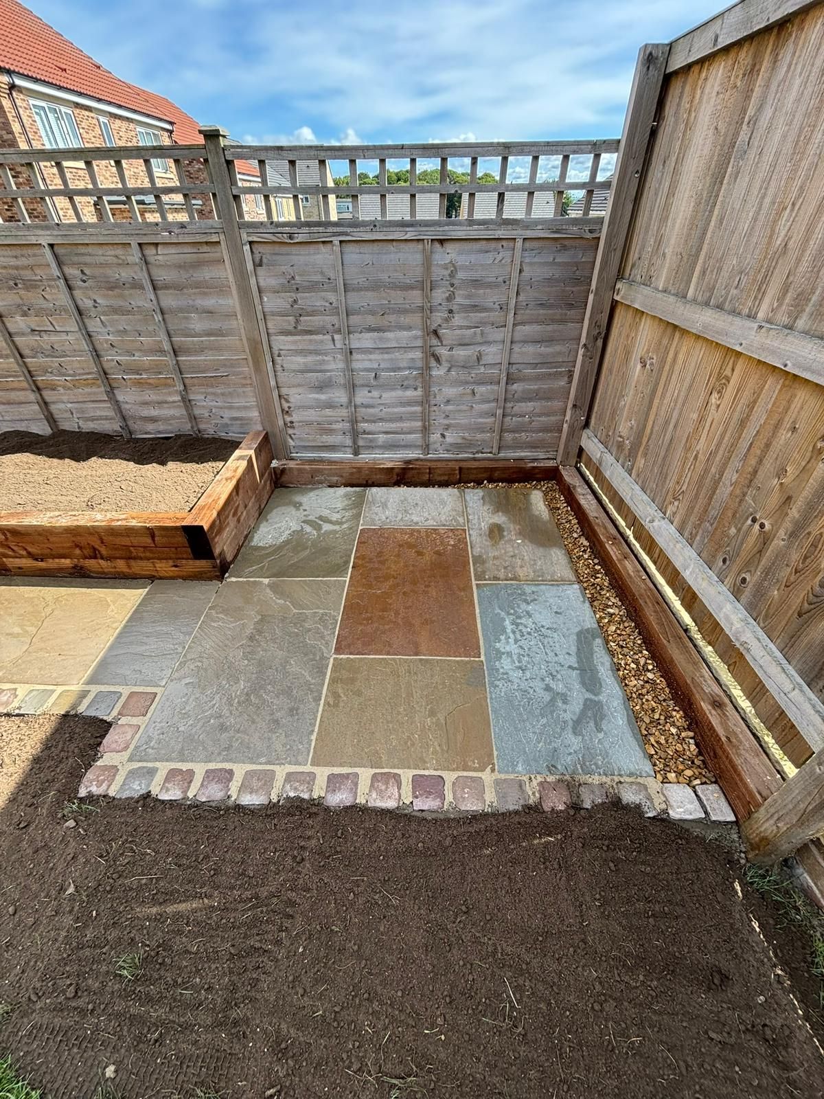 Small fenced backyard patio with square pavers and a soil patch in the foreground.