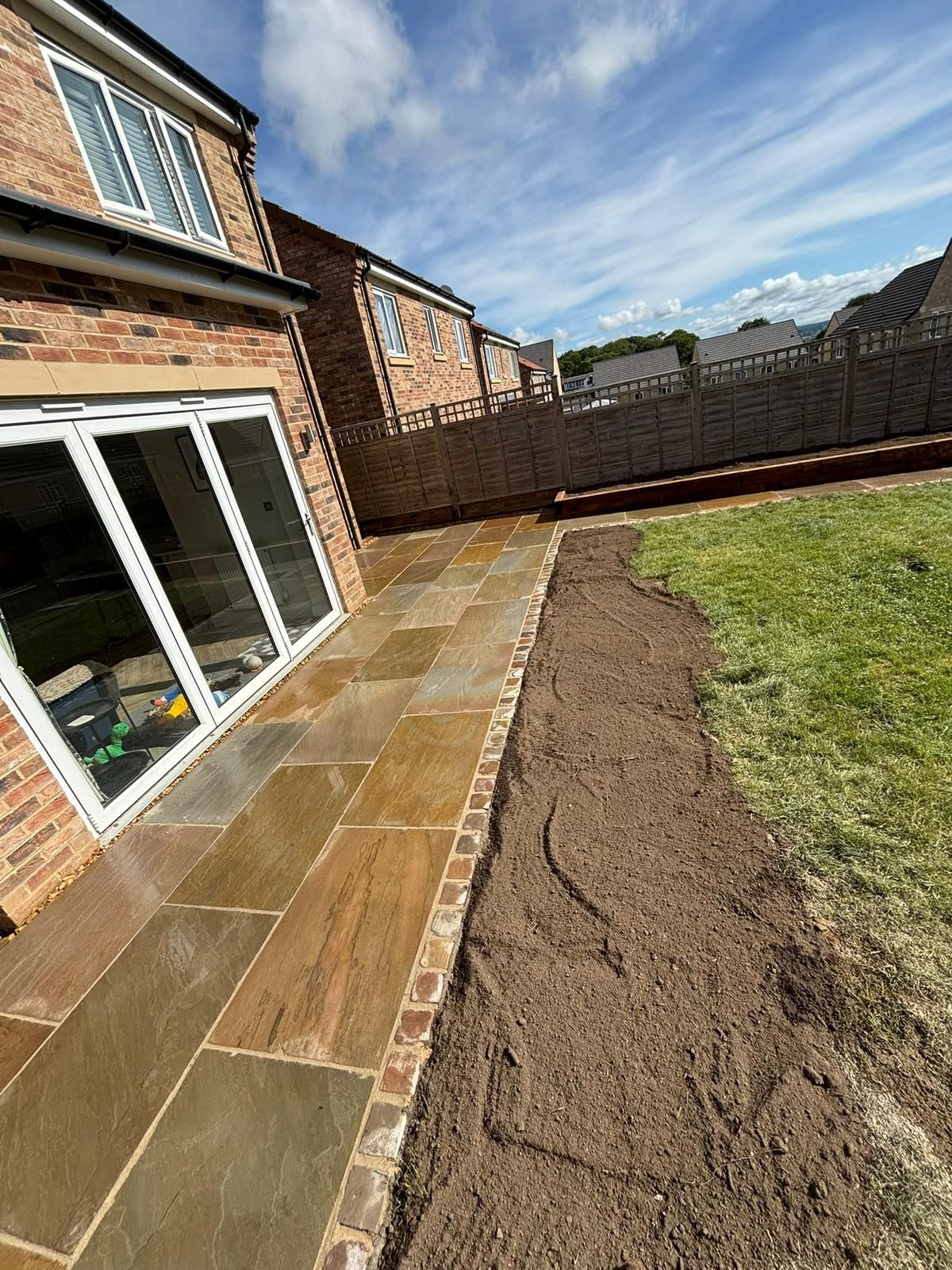 Backyard patio beside a brick house, with new paving slabs, soil border, and lawn under a blue sky.