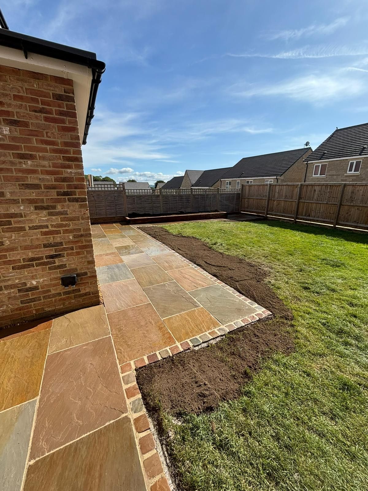 Backyard patio with tan paving beside a brick house, bordered by grass and a wooden fence under a blue sky