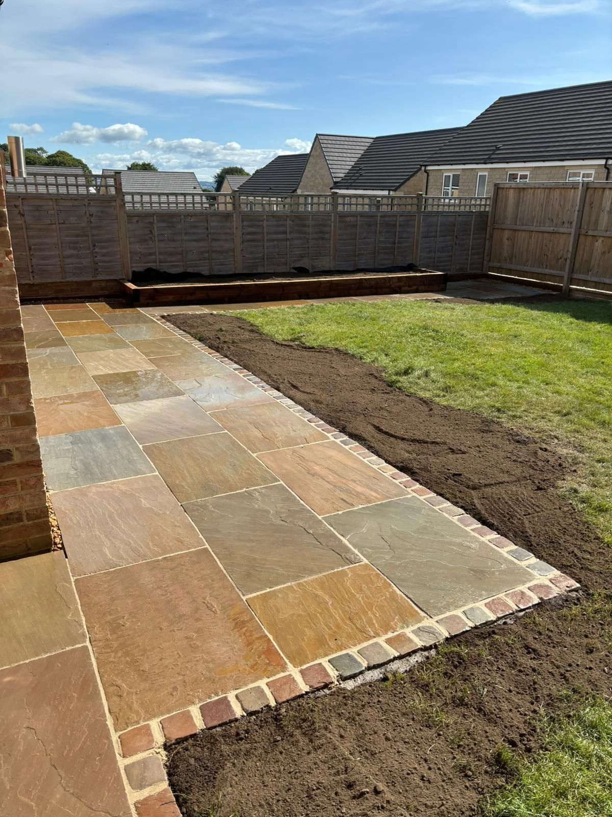 Backyard patio with stone paving, grass lawn, and fenced seating area under a cloudy sky