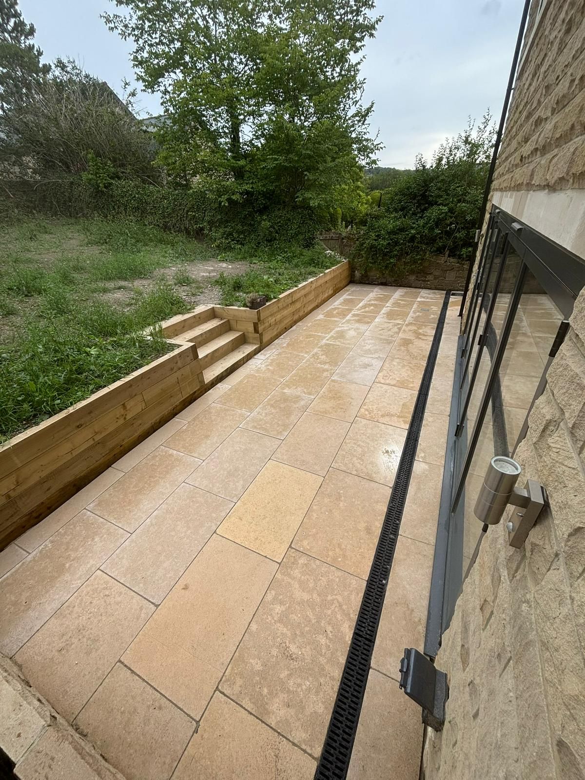 Narrow stone patio beside a house, bordered by wooden planters and steps leading to a grassy yard.