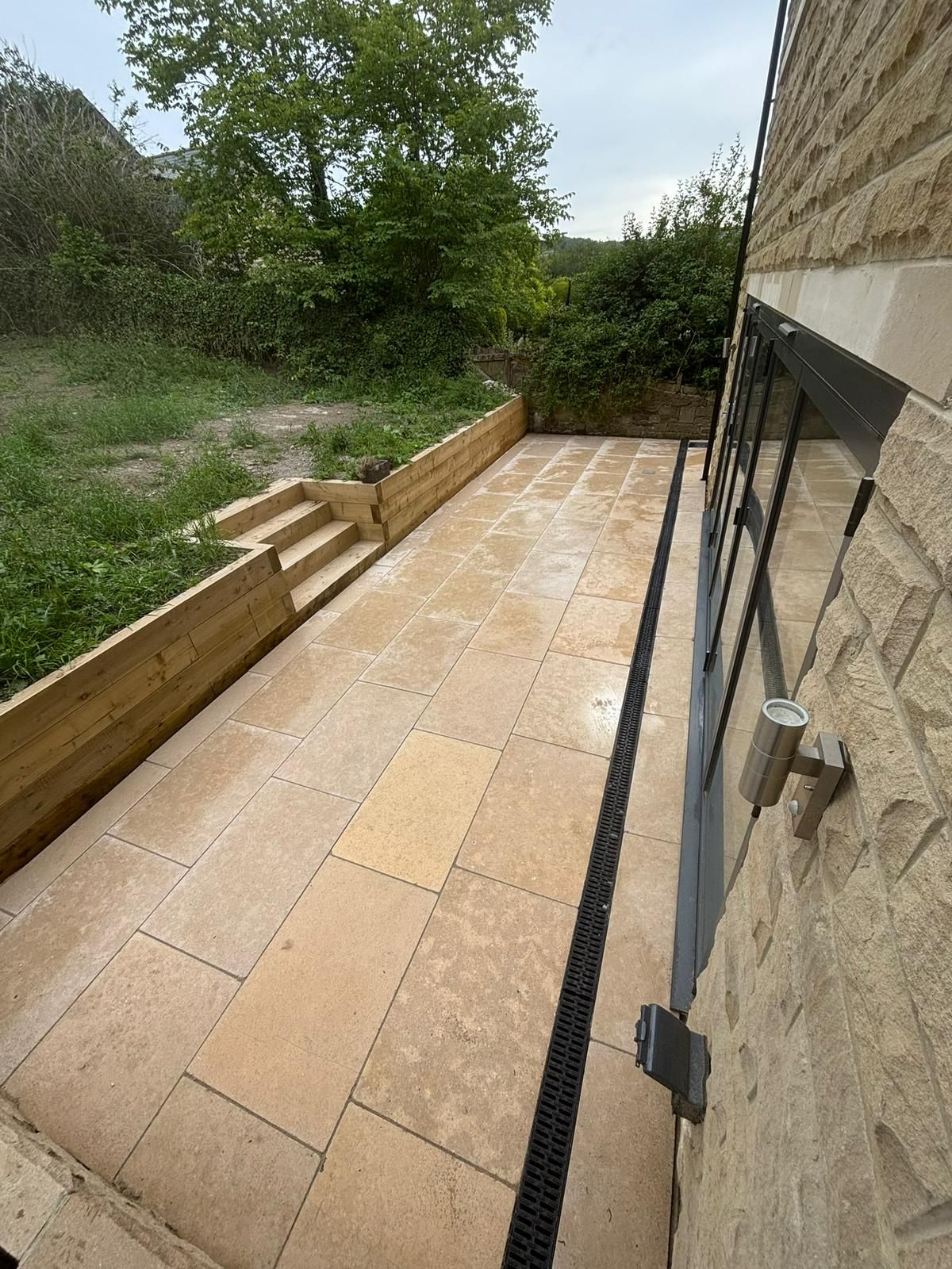Narrow tiled patio beside a stone house, with wooden edging, steps, and greenery at the end.