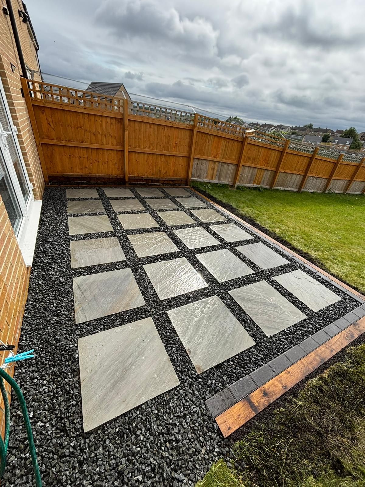 Backyard patio with gray stone pavers set in black gravel beside a wooden fence and grass patch