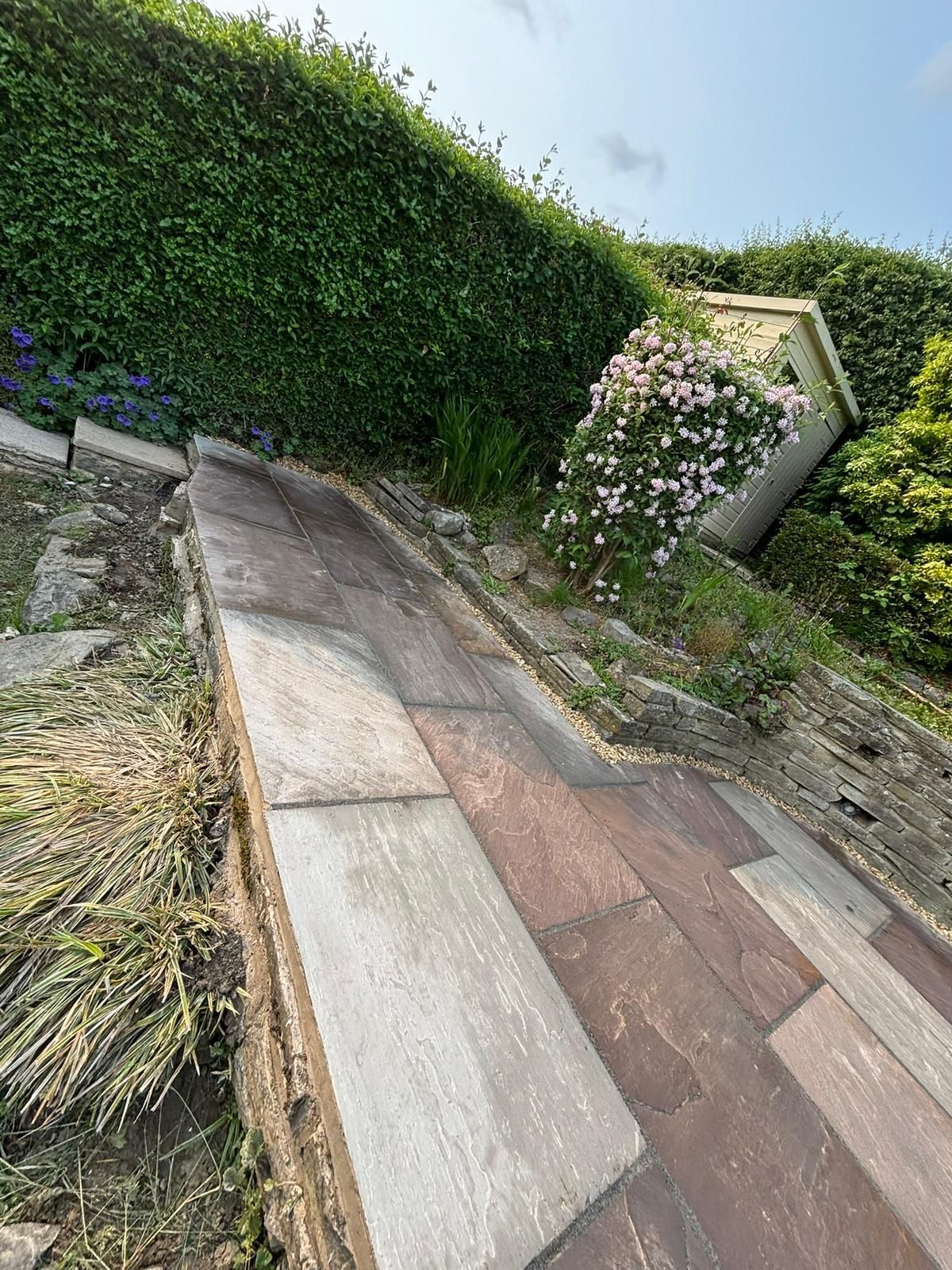 Sloped brick-and-stone walkway beside a hedge and flowering bush under a bright sky