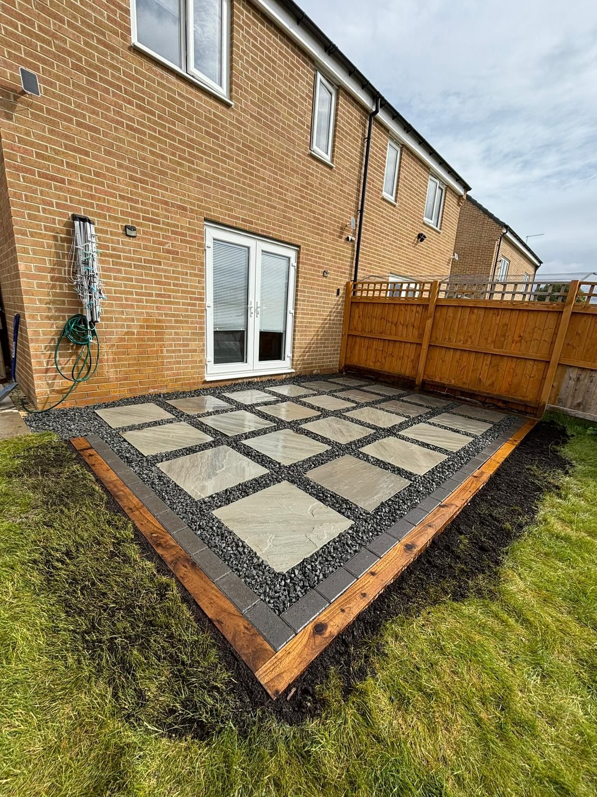 Small brick patio with square pavers beside a house, bordered by lawn and a wooden fence.