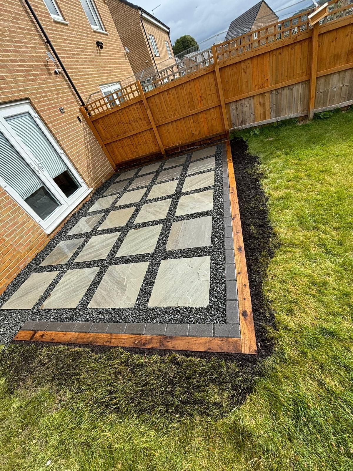 Backyard patio with gray paving slabs, brick border, and wooden fence beside a brick house
