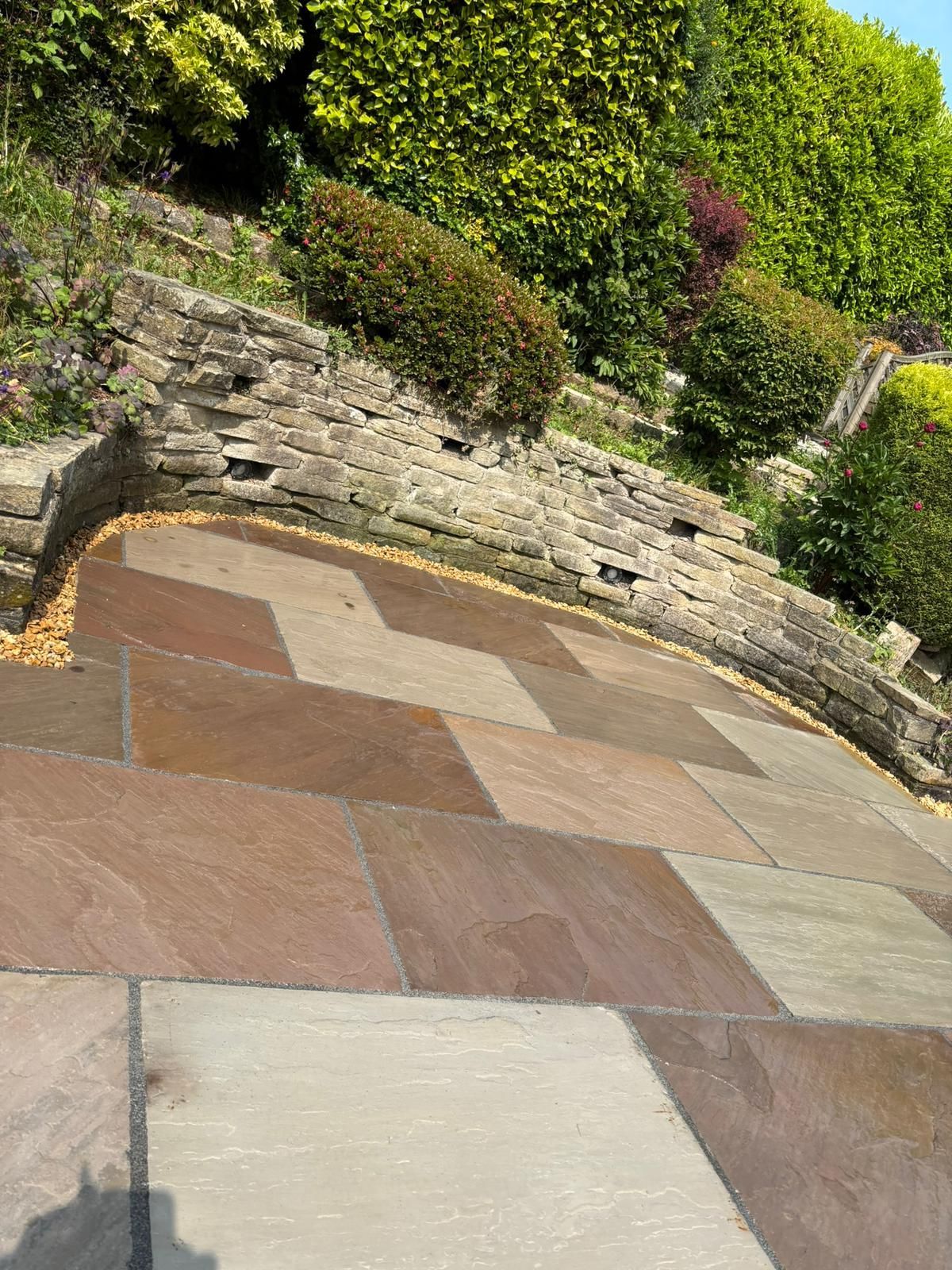 Paved patio with beige and brown stone tiles beside a low stone wall and greenery on a sloped garden.