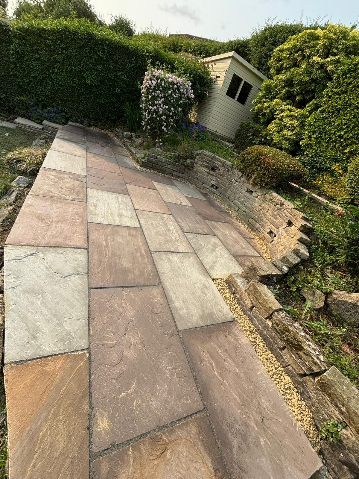 Stone patio path beside a trimmed hedge and small garden shed on a sunny day