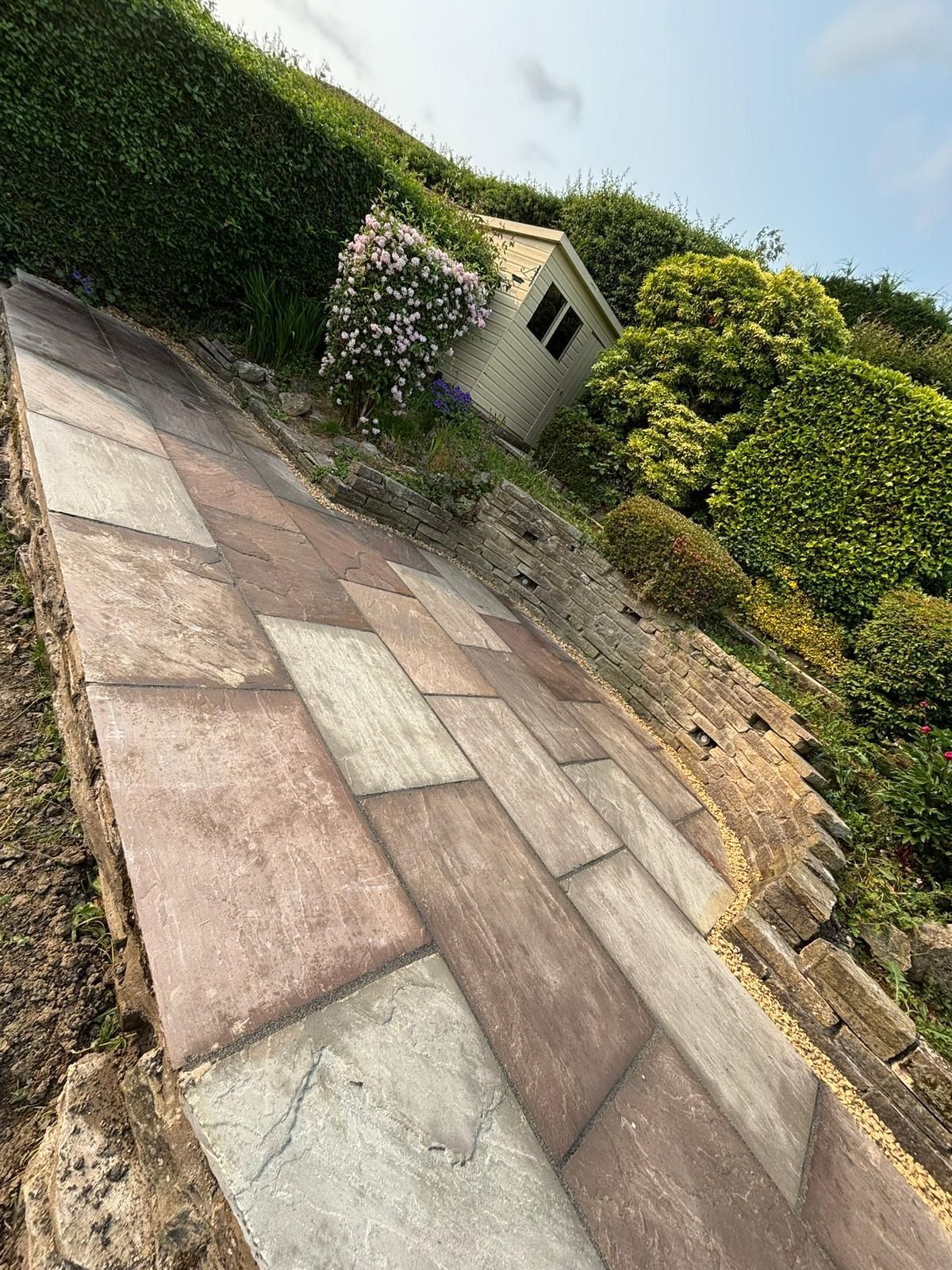 Sloped stone patio beside tall hedges and a small garden shed under a bright sky