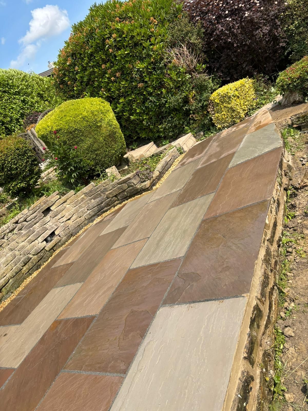 Slanted stone garden path bordered by shrubs and a low stone wall under a blue sky