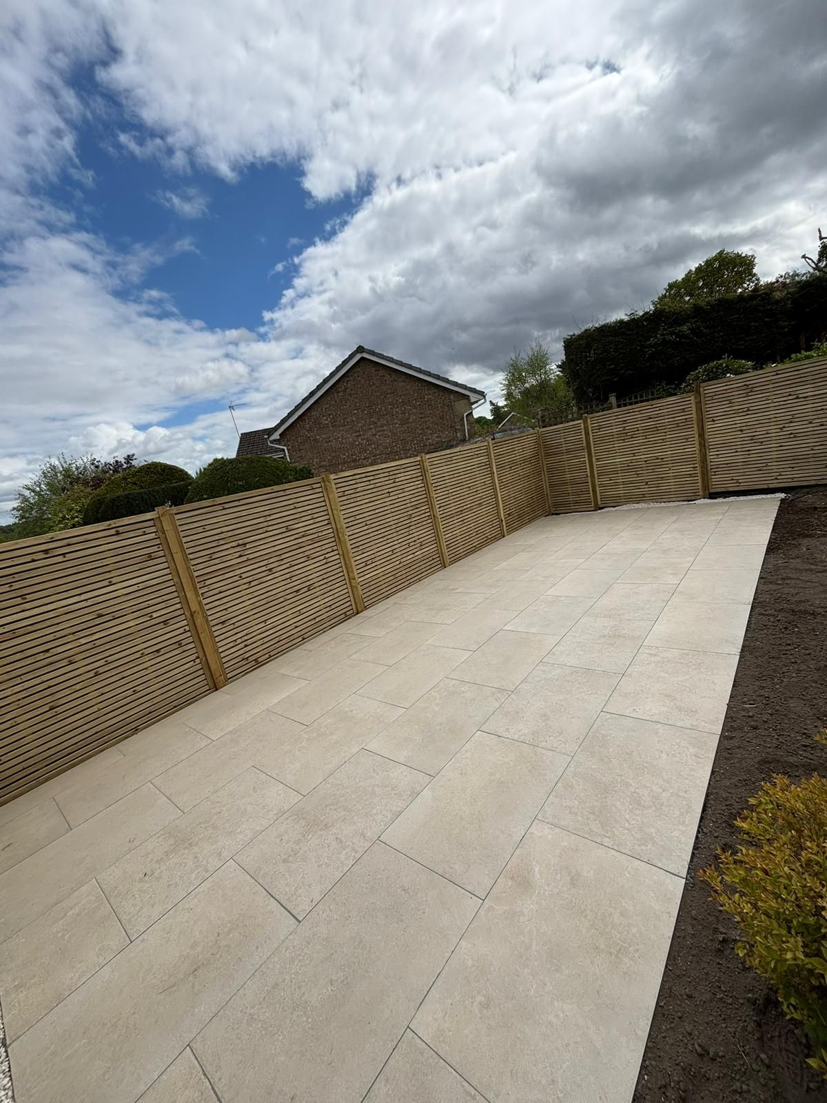 Wide concrete driveway beside a wooden fence, sloping upward under a cloudy sky.