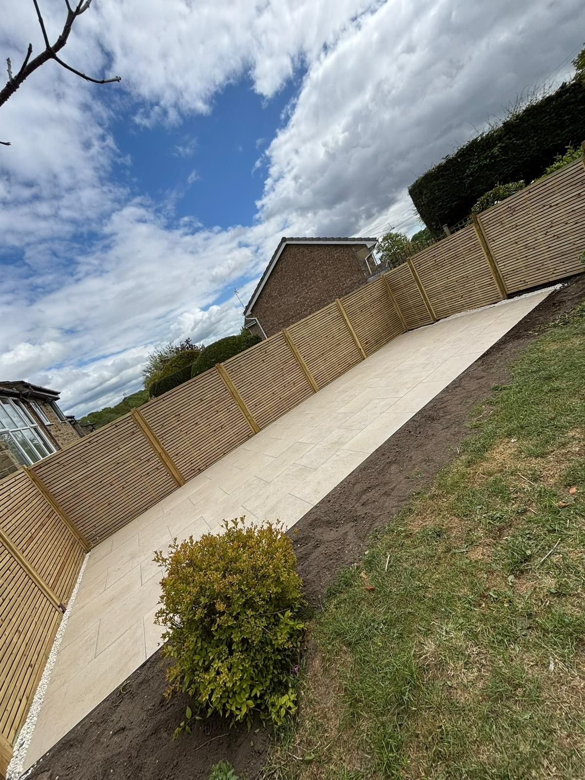 Slanted concrete driveway beside a wooden fence, with a small bush and cloudy blue sky overhead
