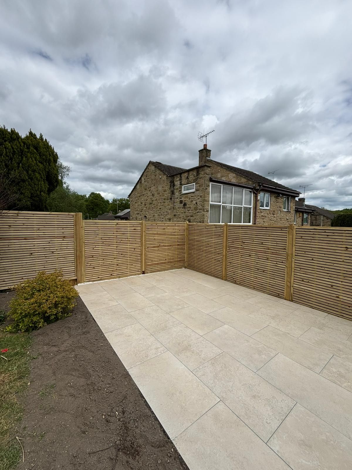 Backyard patio with pale paving stones, timber fence, and a stone house under cloudy sky