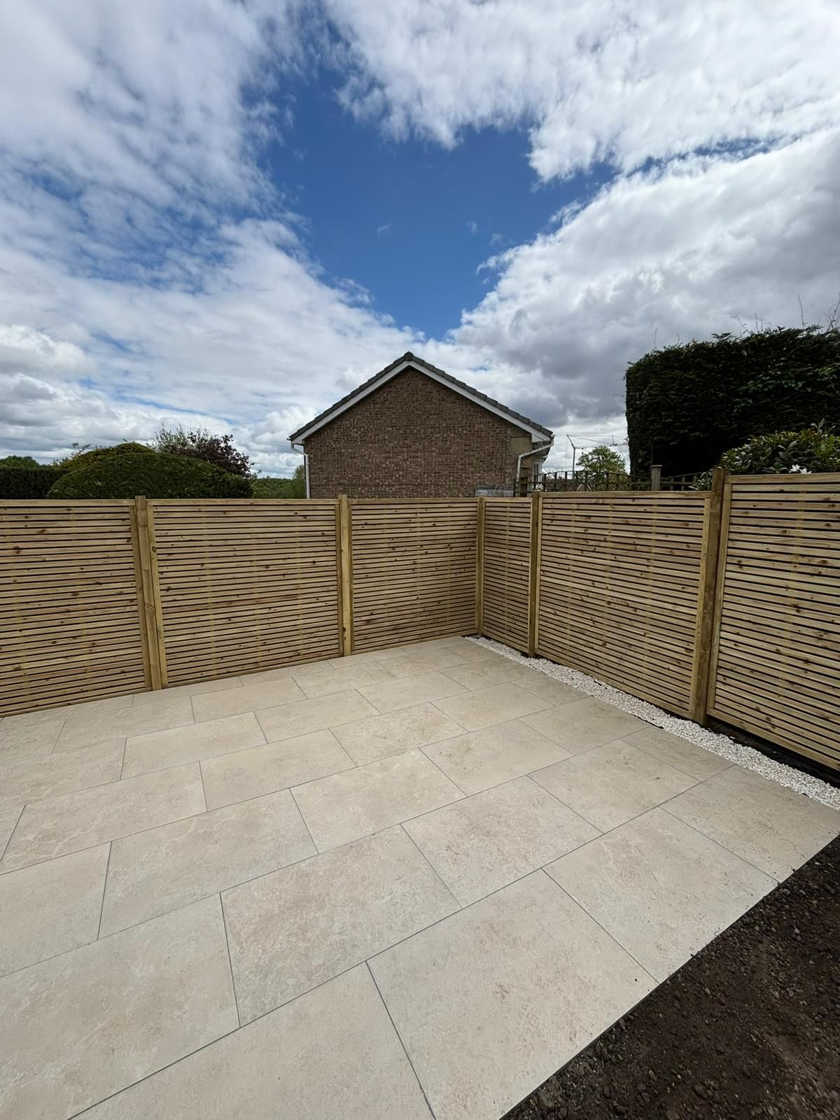 Empty paved patio with beige fence panels and a brick house under a cloudy sky.