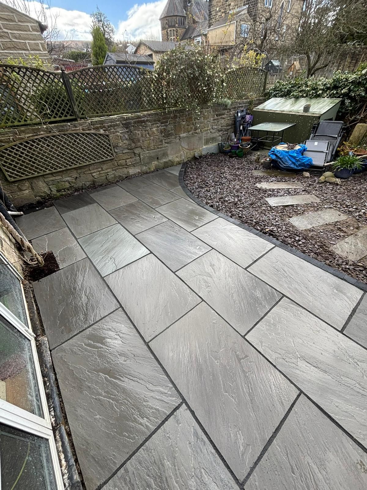 Small paved patio with wet gray tiles, gravel border, and a fenced garden area with outdoor items.