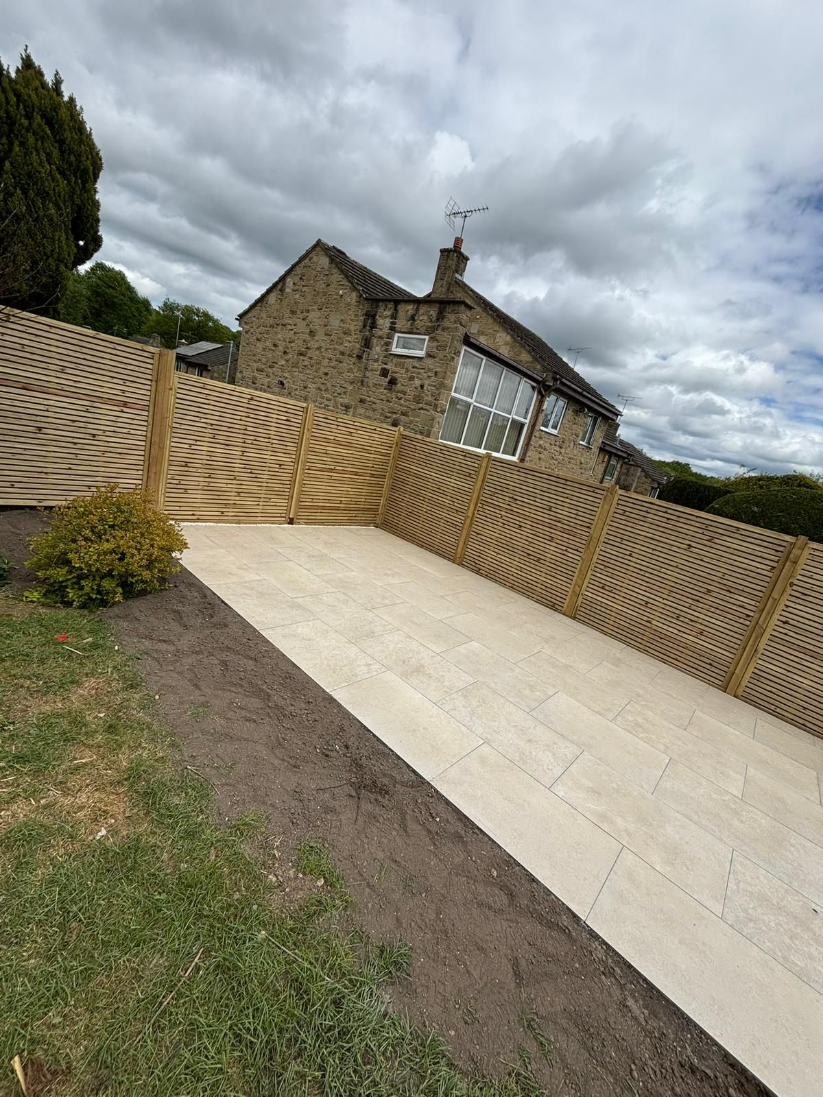 Newly paved driveway beside a stone house under a cloudy sky, with wooden fencing on both sides