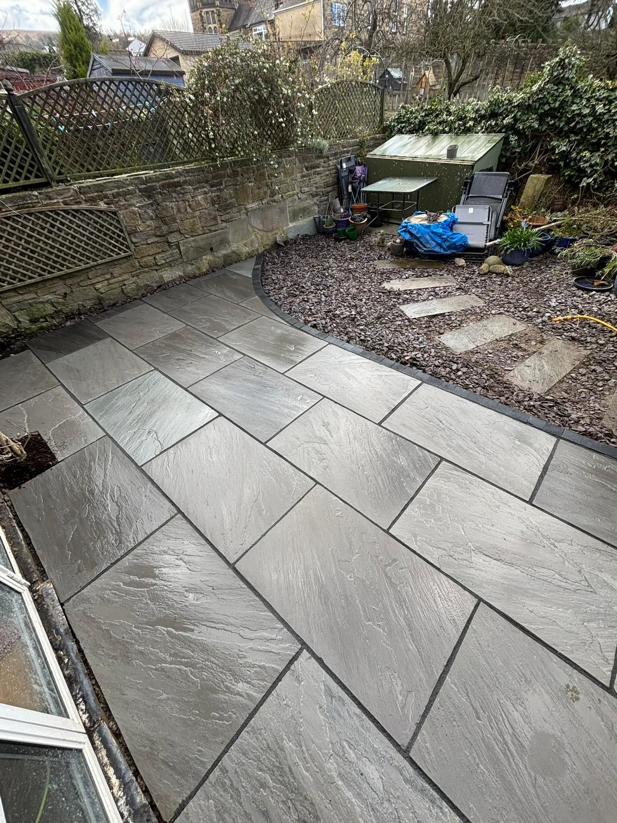 Wet gray patio tiles beside a gravel garden, with a fence, shed, and blue tarp in the background