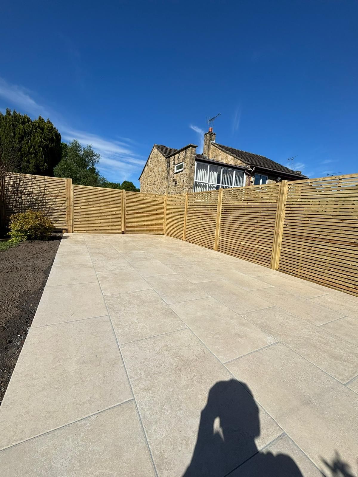 Wide stone driveway beside a wooden fence under a bright blue sky, with a house in the background.