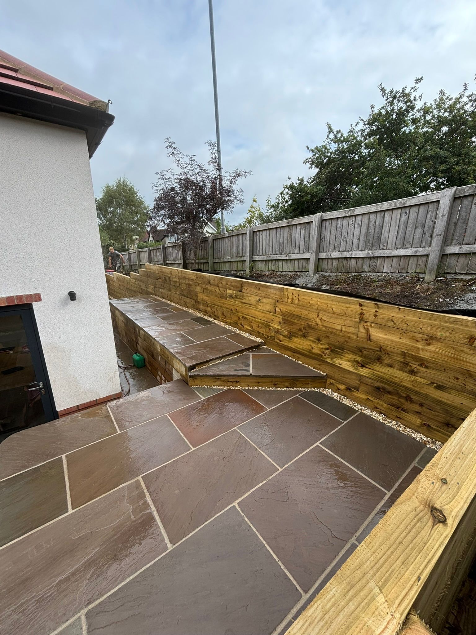 Rain-soaked tiled patio beside a white house, with wooden retaining wall and fence under a cloudy sky