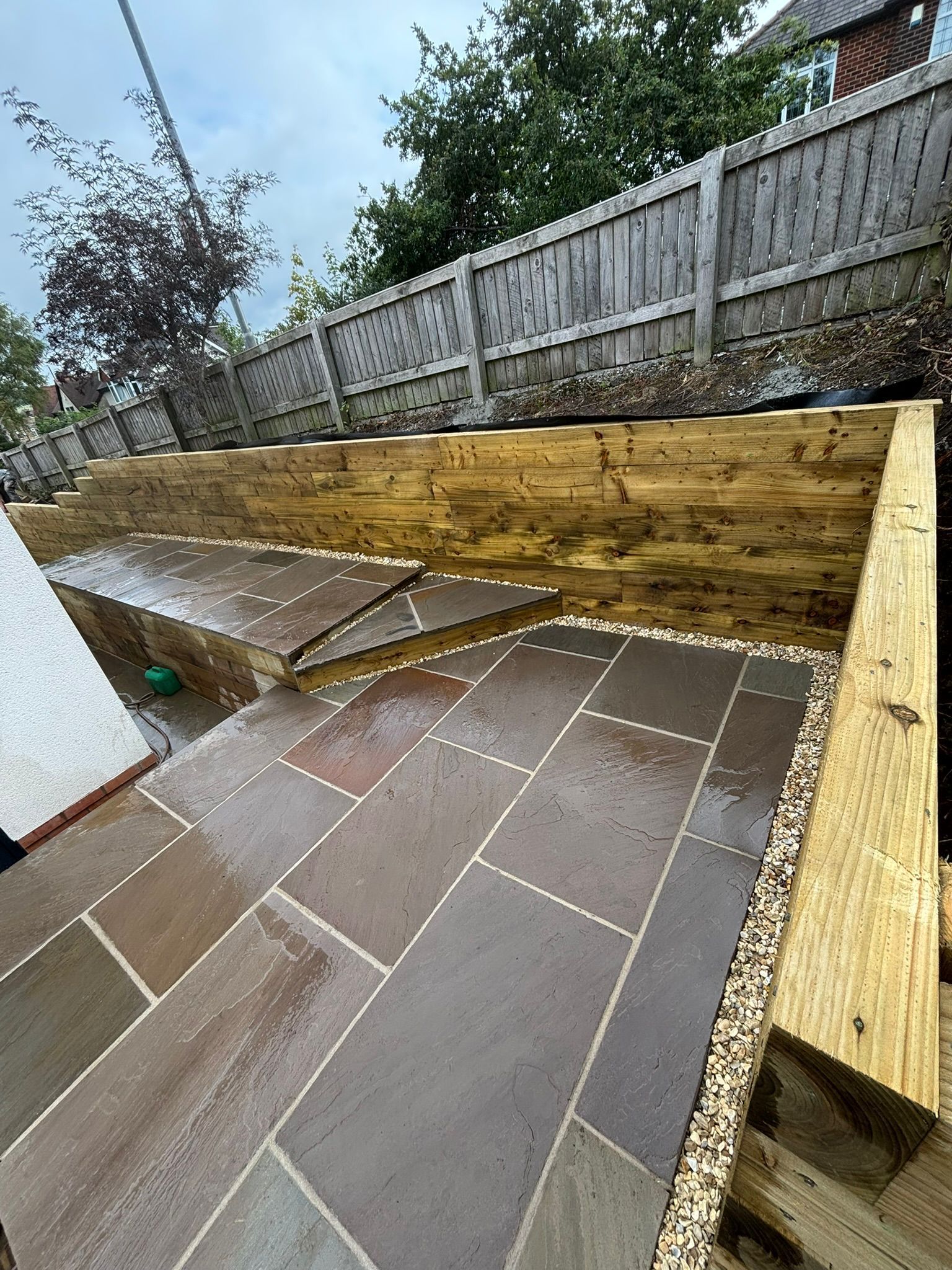 Wooden retaining wall around a wet tiled patio beside a fenced backyard