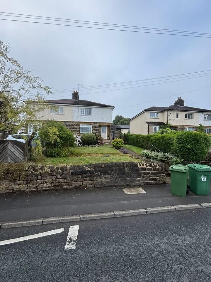 Residential houses with front gardens, stone wall, and green bins along a street under a cloudy sky