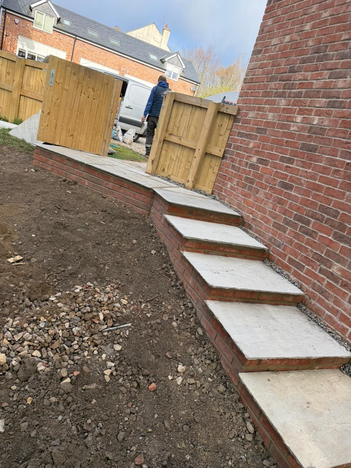 Brick wall beside a dirt yard with concrete stepping stones leading to a wooden gate