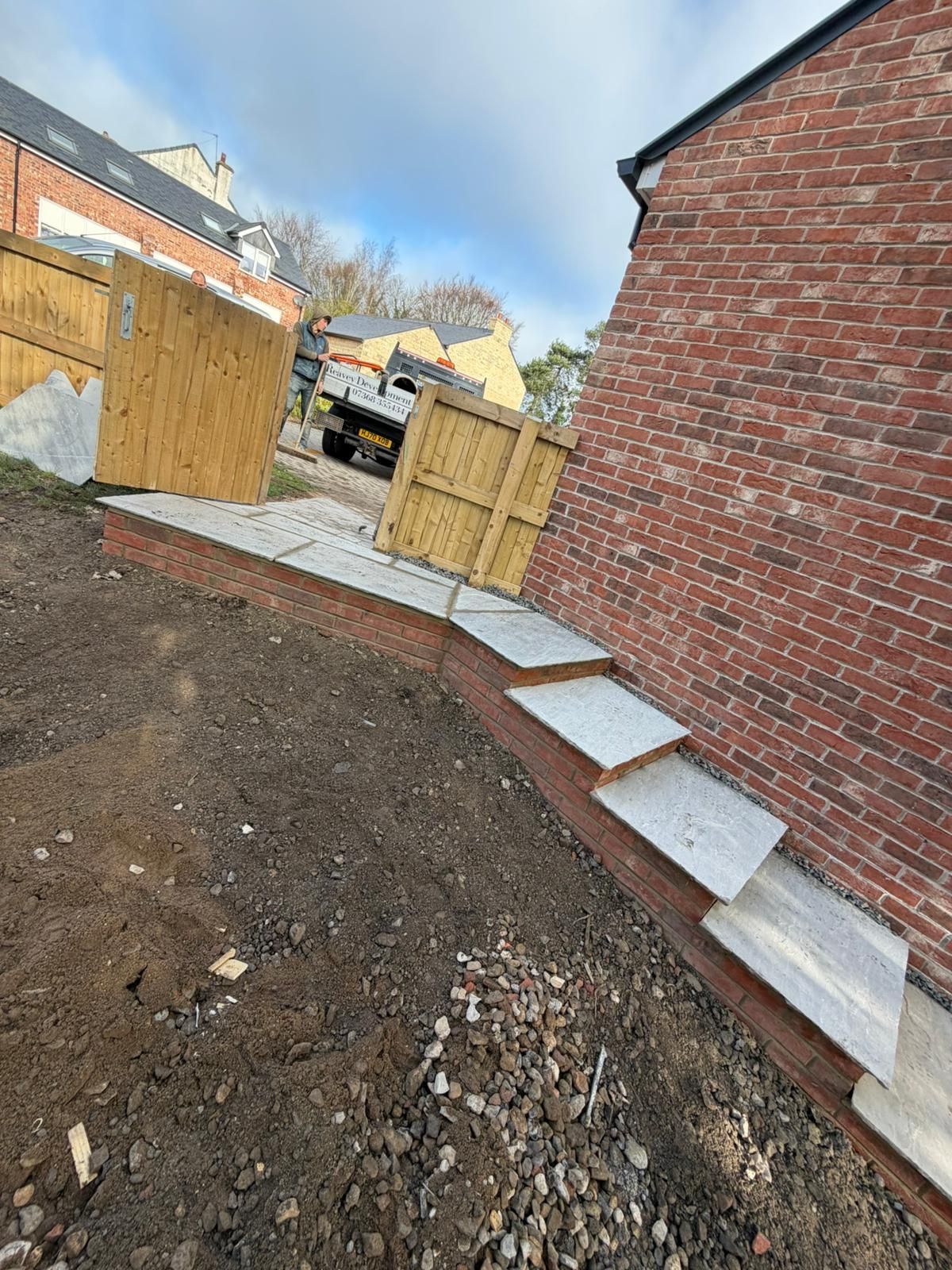 Backyard construction area with dirt, wooden fence panels, and a brick house wall along a paved edge