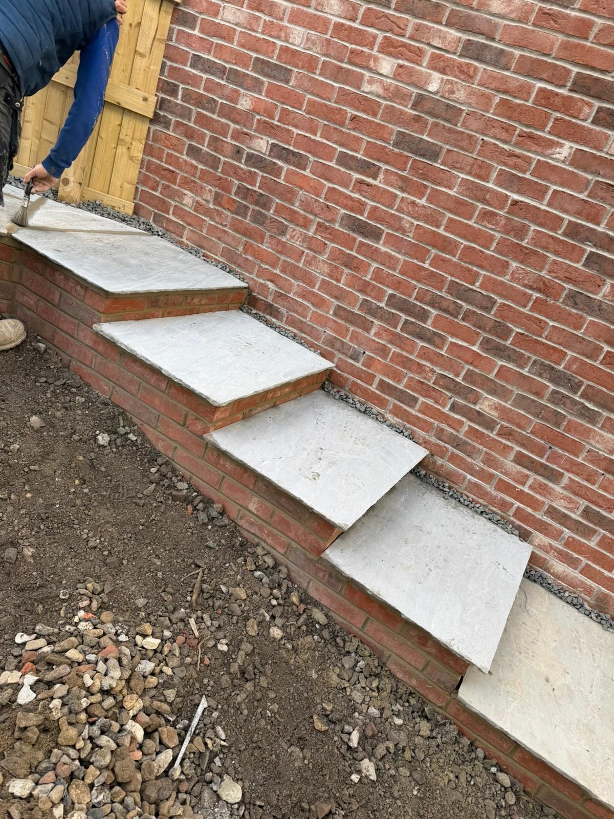 Worker installing white stone steps beside a red brick wall on a dirt slope