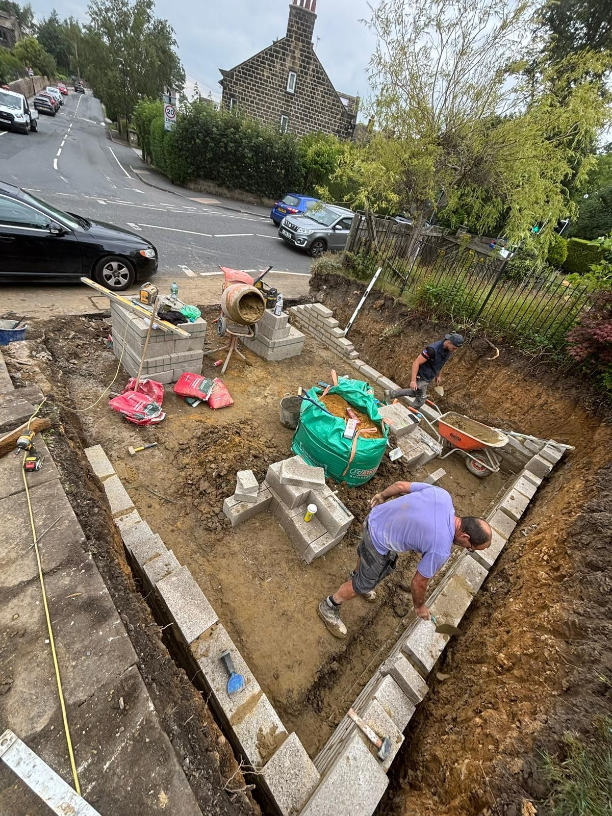 Garden bed with two workers and bags of soil beside a street, near a parked car and stone house