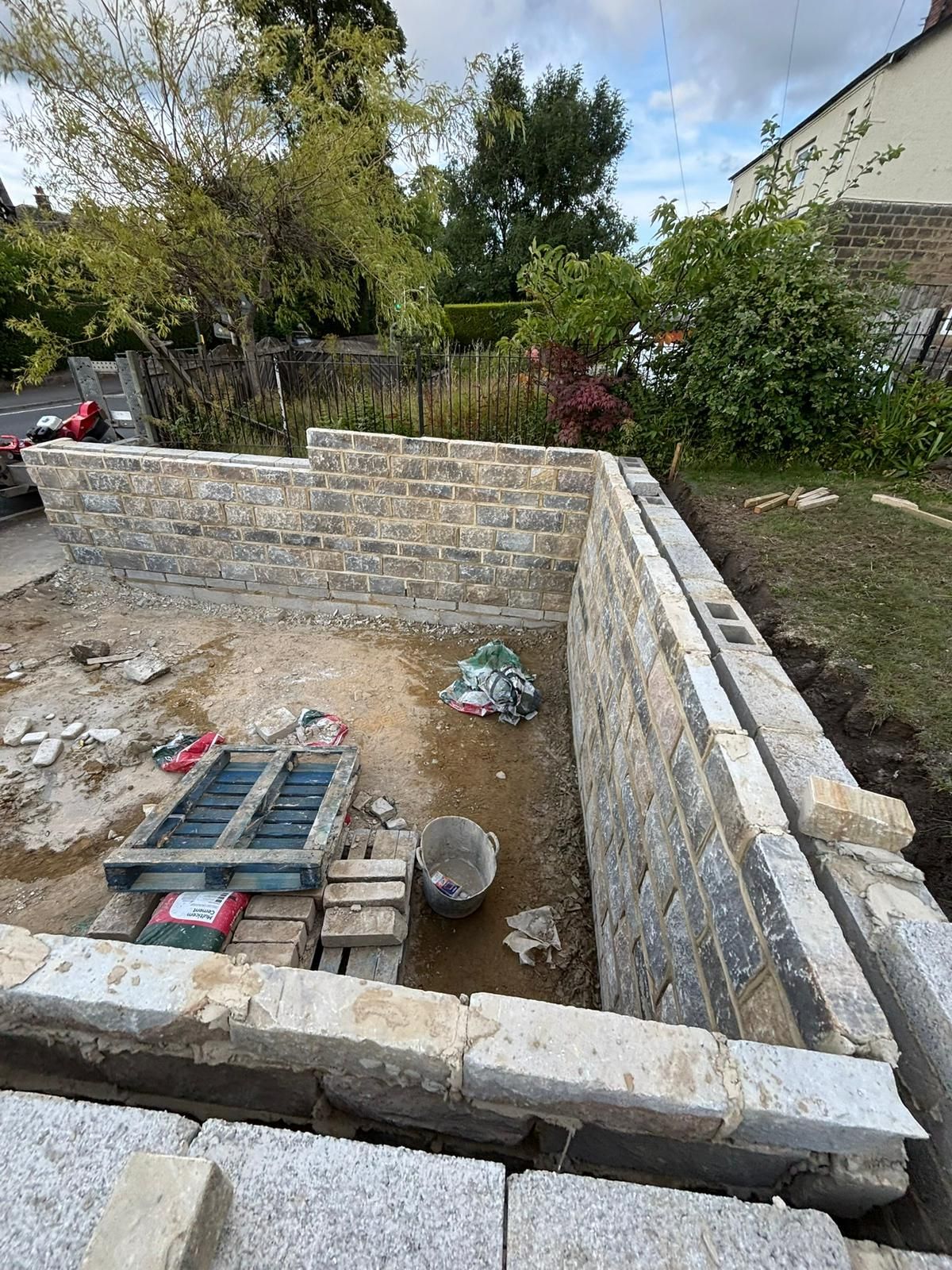 Stone foundation under construction in a backyard, with stacked bricks and tools on the ground.