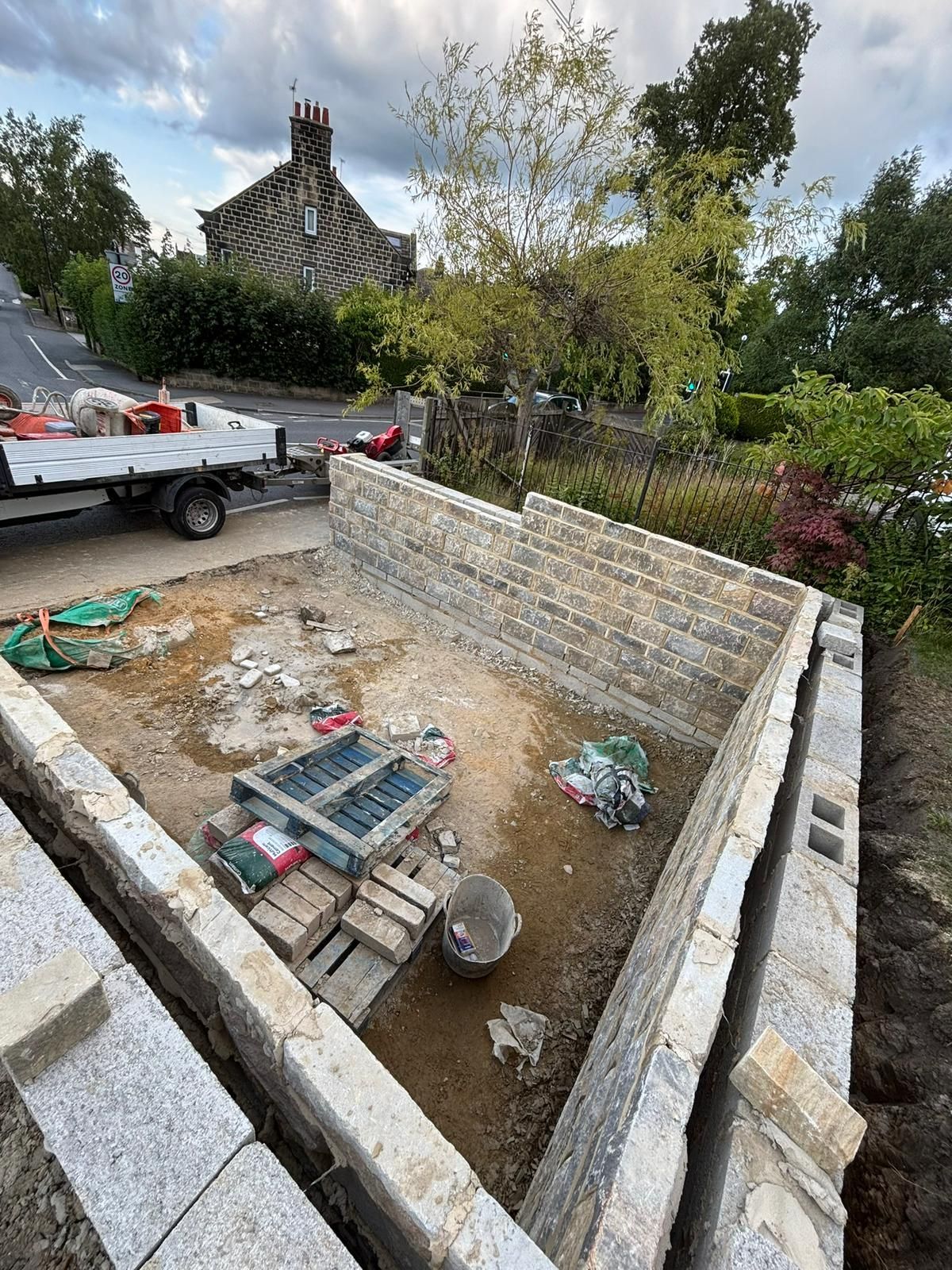 Partially built stone foundation with pallets and debris in a yard, near a trailer and trees
