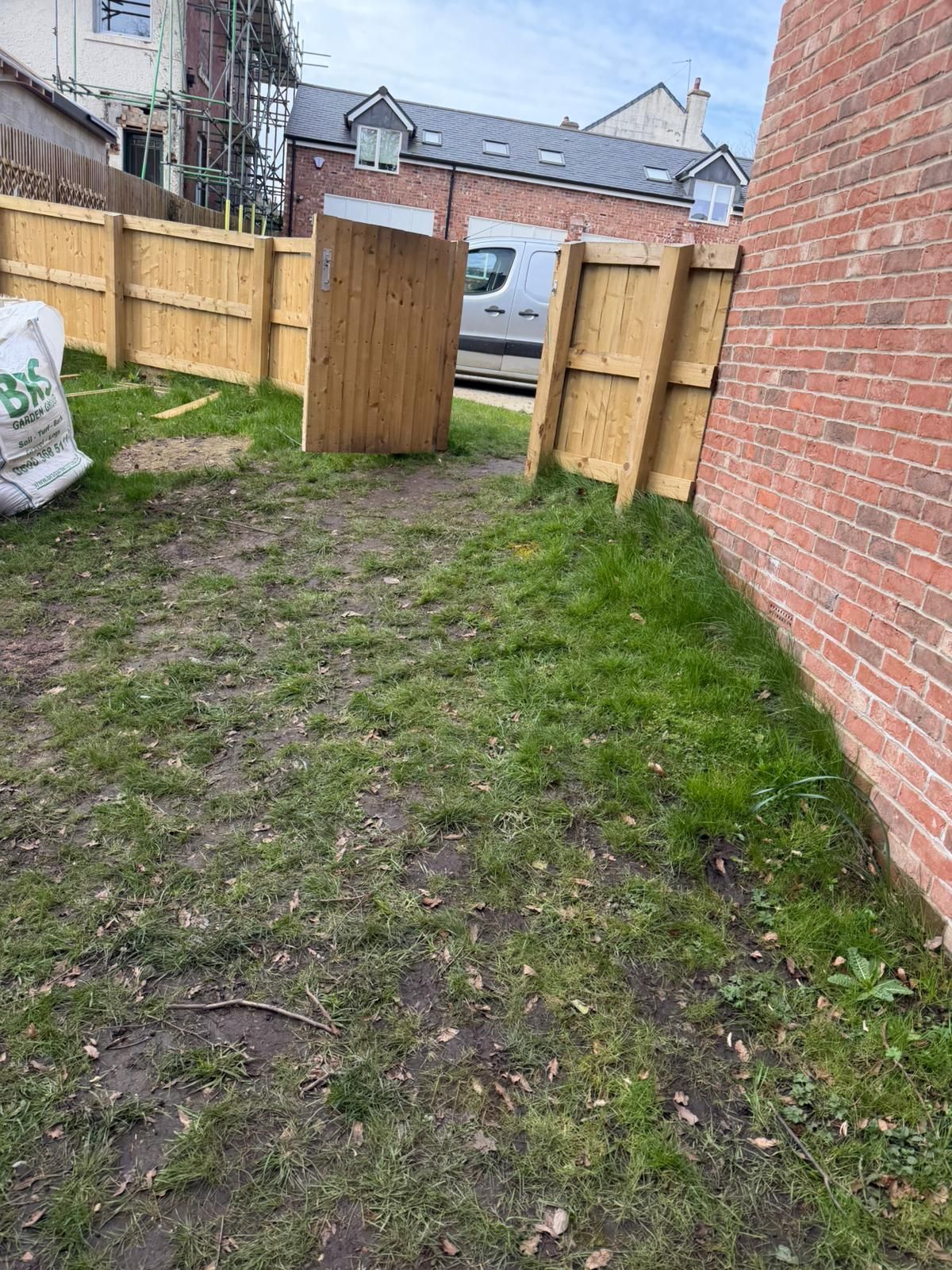 Narrow grassy backyard path beside a brick house and wooden fence with an open gate and shed beyond.