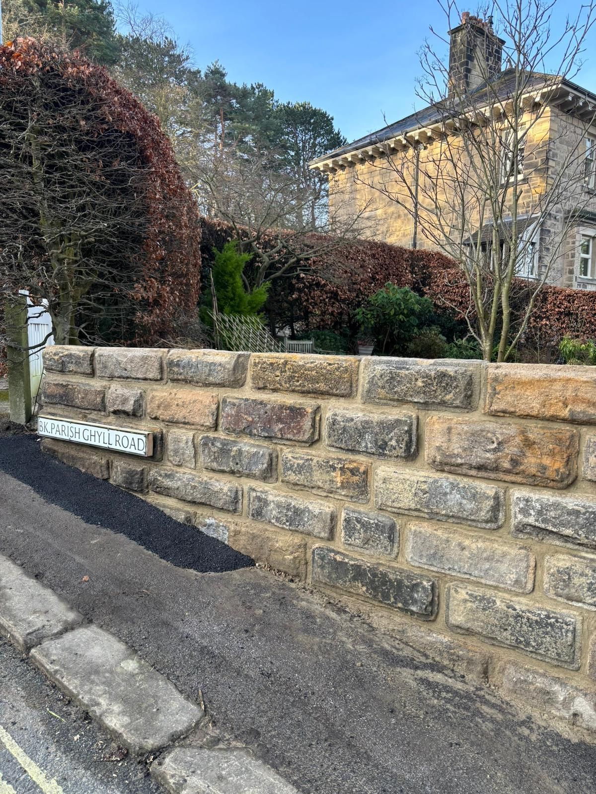 Stone retaining wall beside a sidewalk, with hedges and a yellow house in the background.