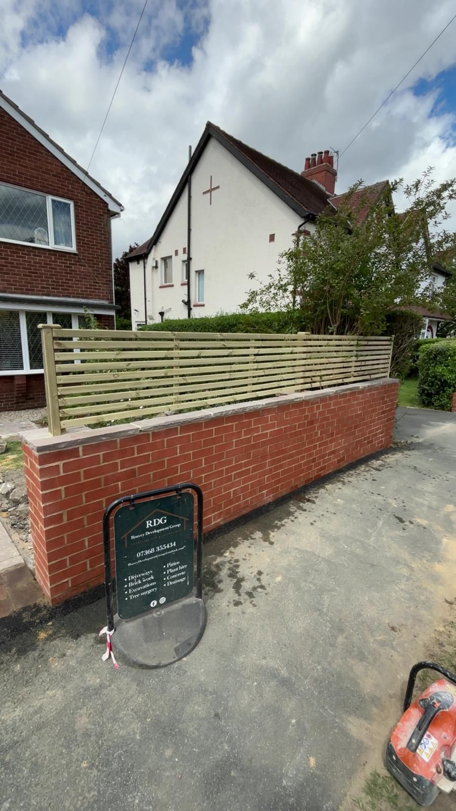 Residential driveway with low brick wall, wooden fence, and a house under a cloudy sky