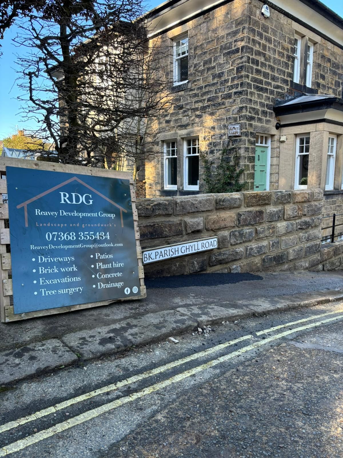 Blue RXC sign outside a stone building on a street corner, beside a low wall and leafless tree.